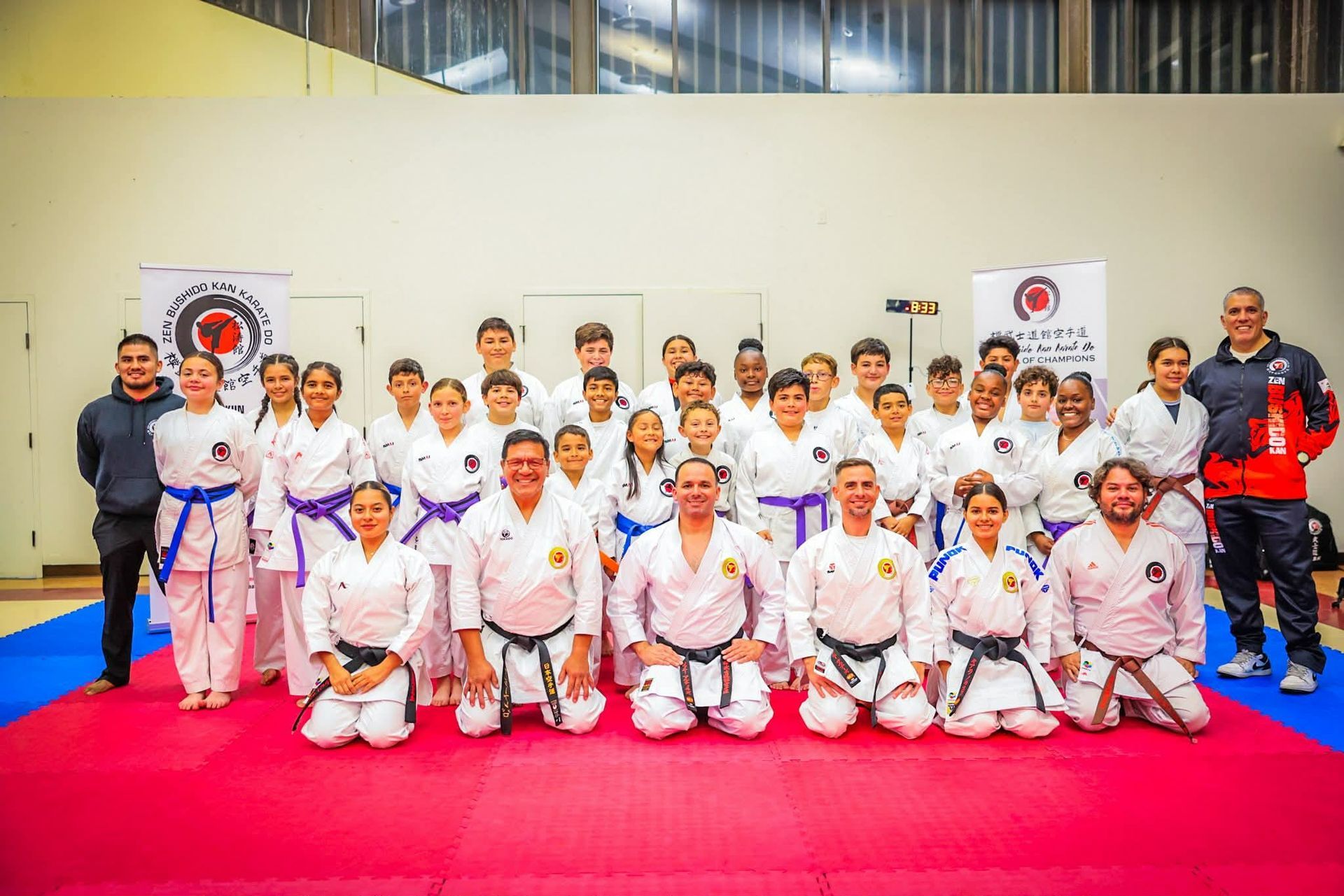 Group of karate practitioners in white uniforms with belts, posing for a group photo in a gym.
