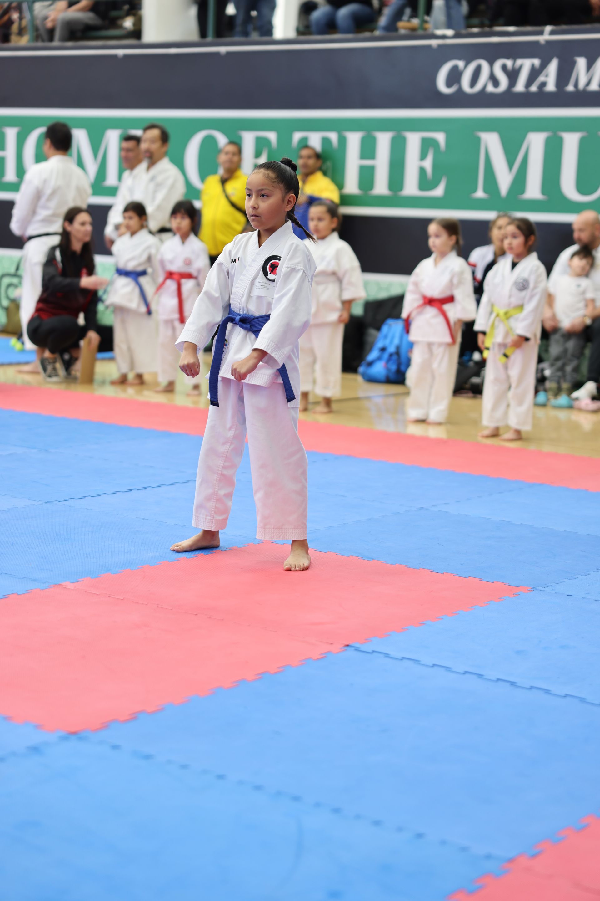 Young karateka in white uniform and blue belt, standing on a blue and red mat in a competition hall.
