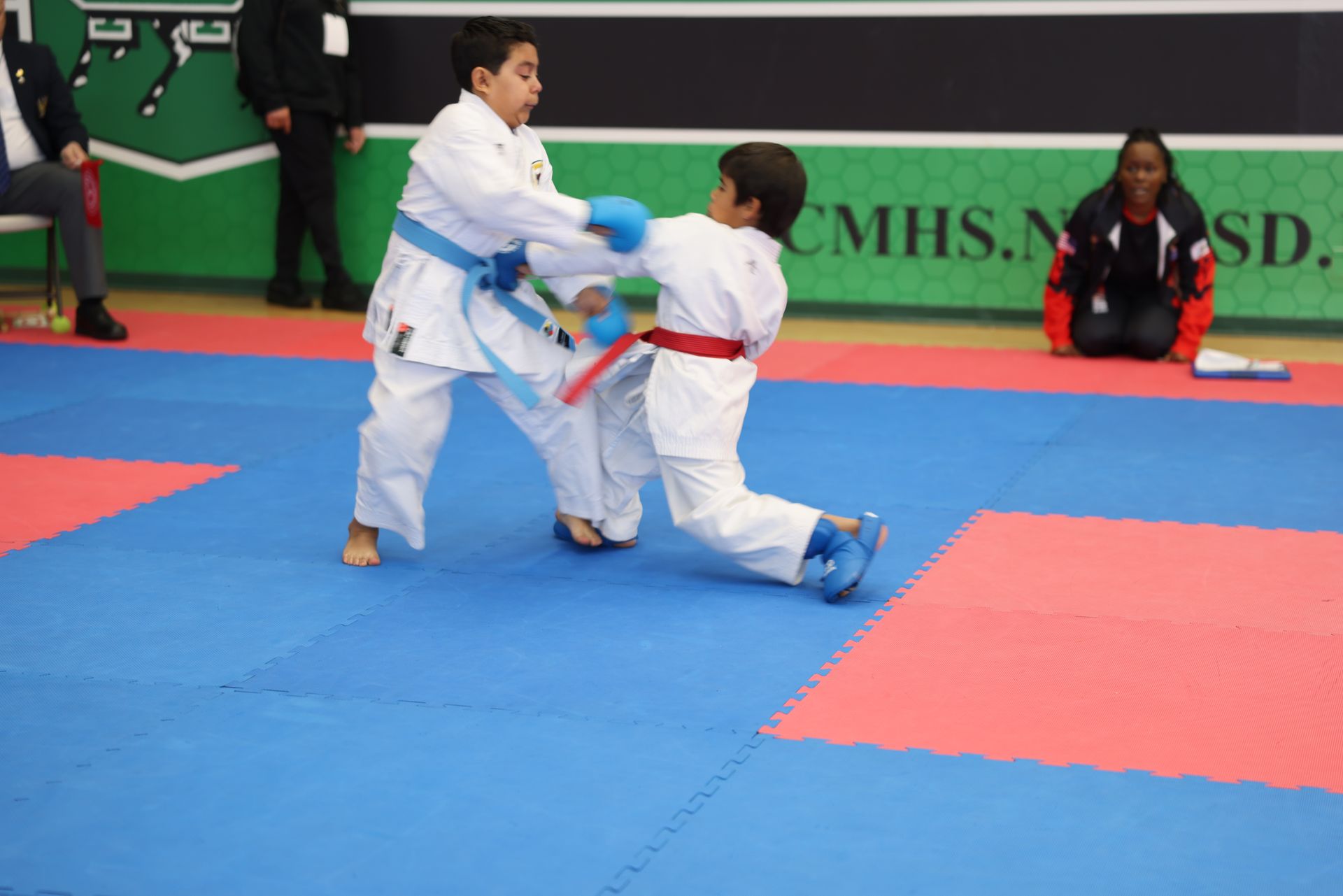 Two karate students sparring on a blue and red mat; one throws a punch, the other ducks.