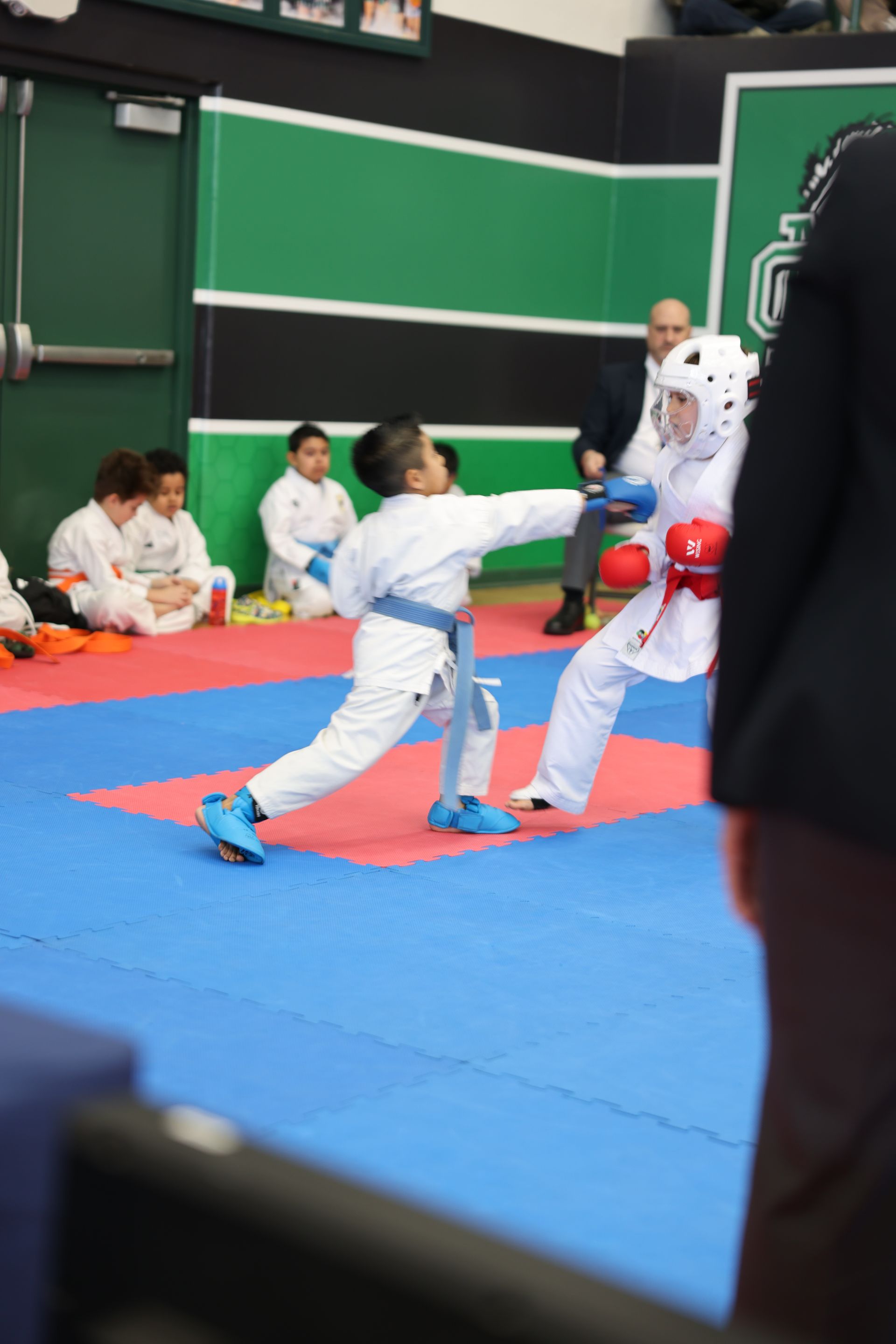 Two karate competitors sparring on a blue and red mat. One throws a punch; the other wears protective gear.