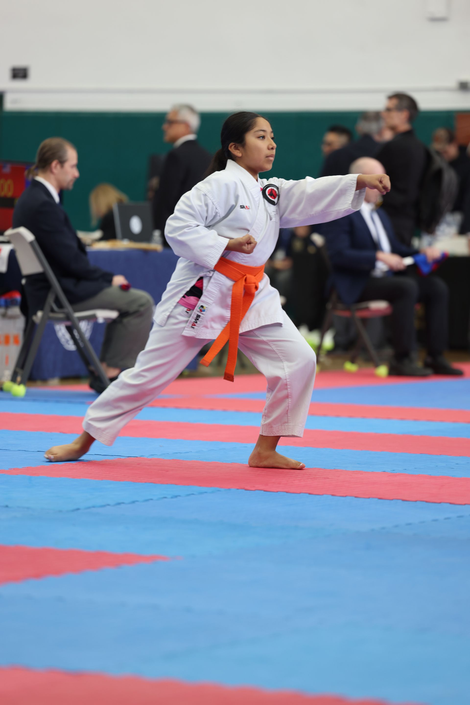 Karateka in orange belt performing a punch during a competition on a blue and red mat.