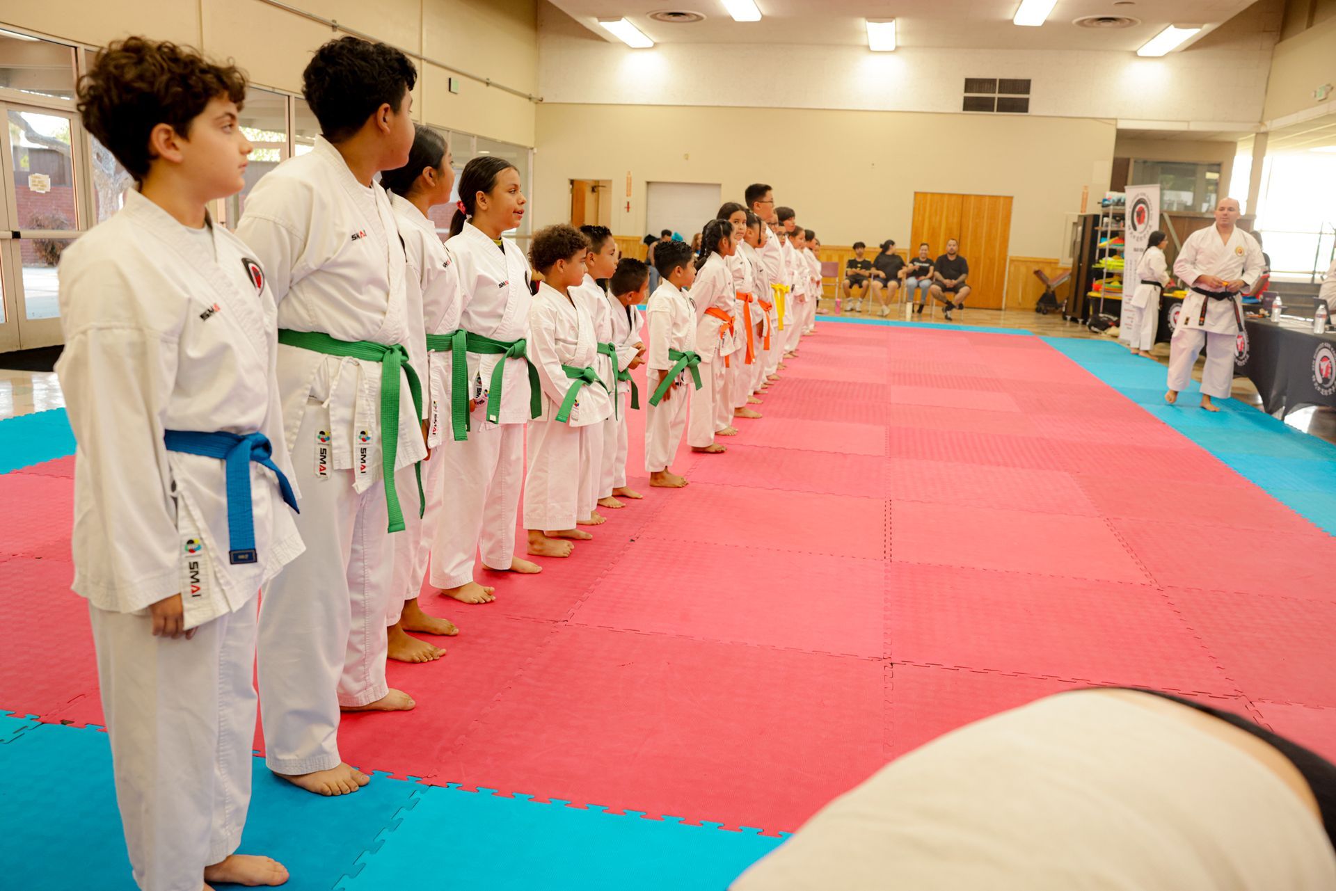 Martial arts students in white uniforms line up on mats in a dojo