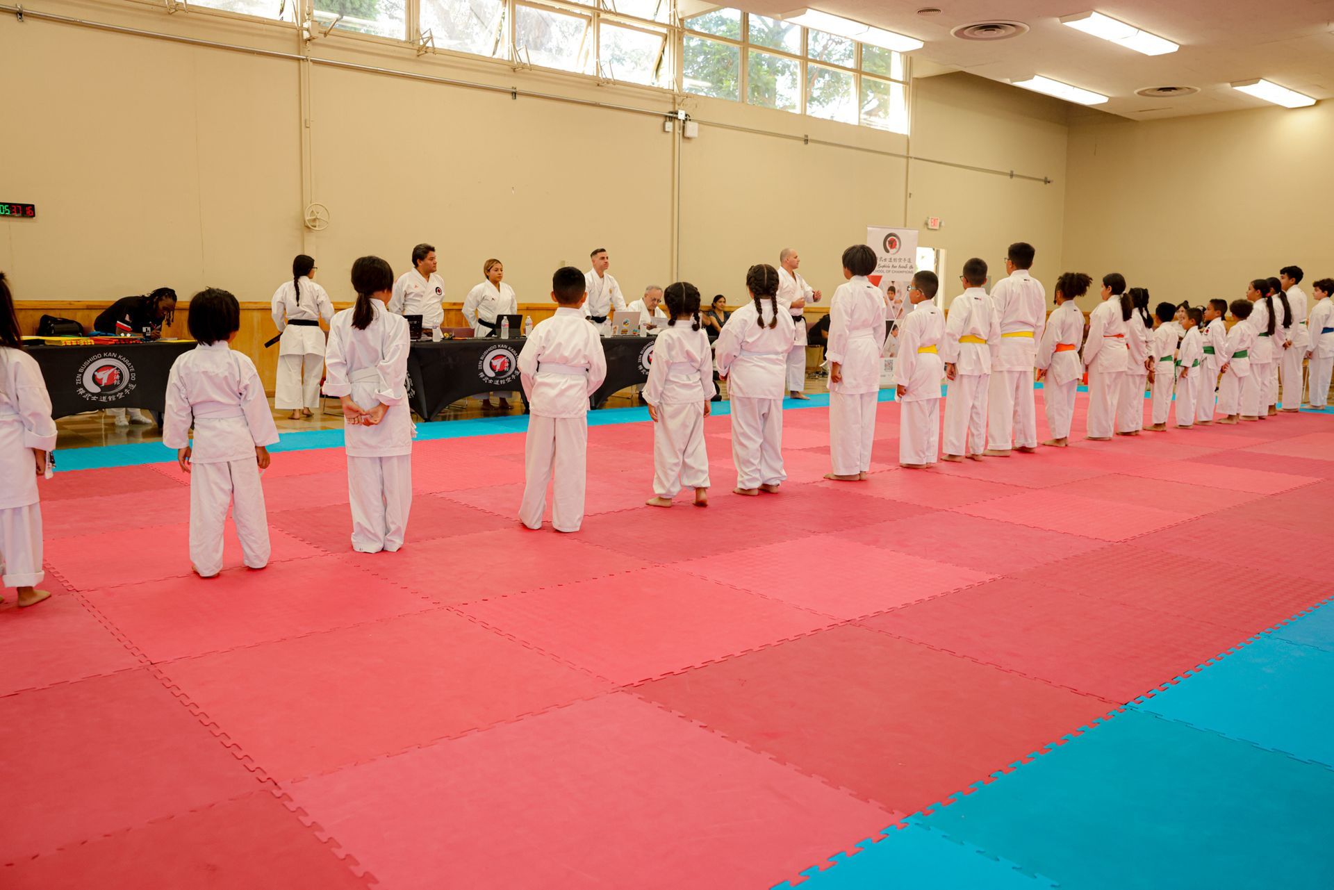 Children in white martial arts uniforms standing in rows on a  training mat in a gymnasium