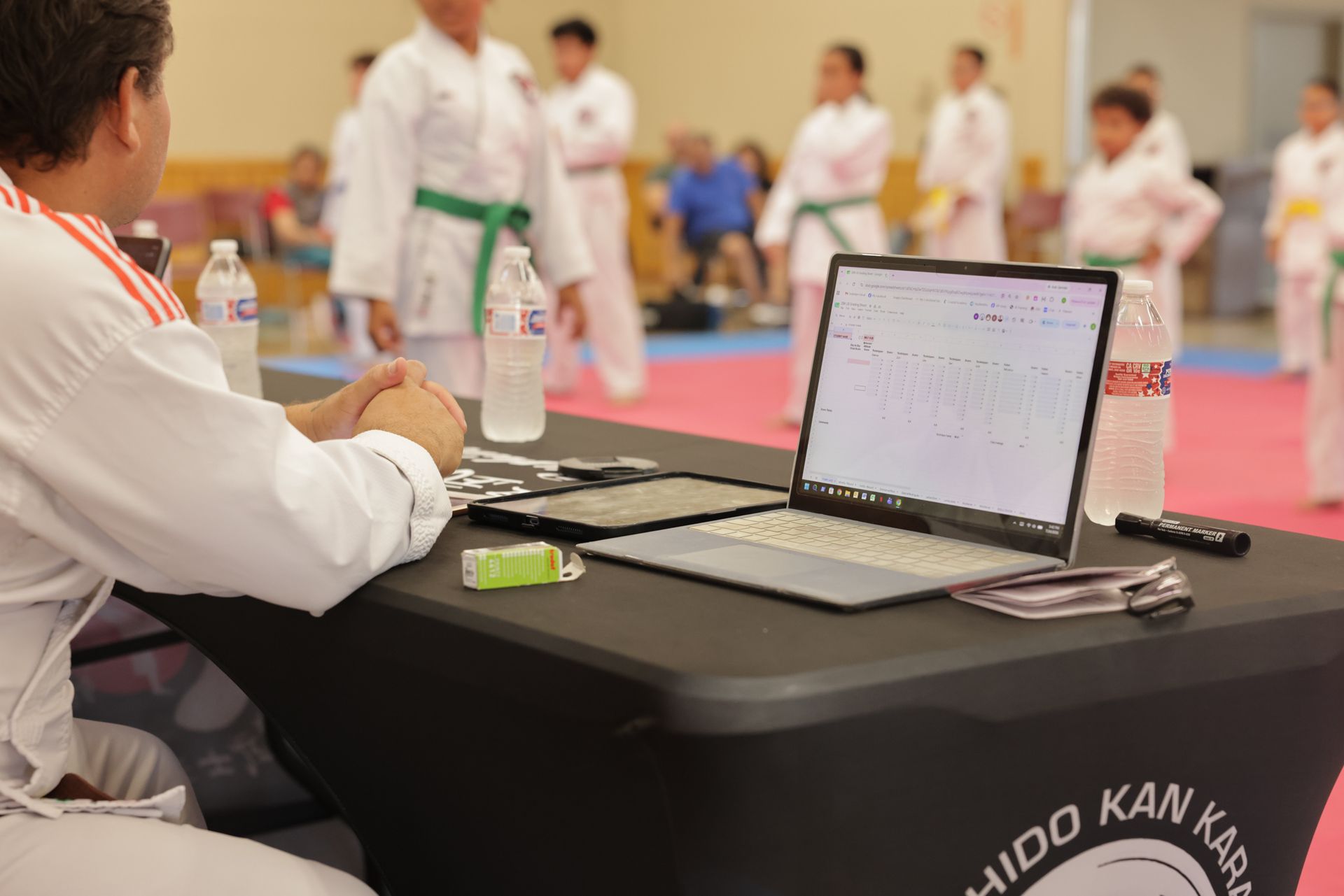 Karate referee table with laptop and papers beside a tatami mat during a martial arts event