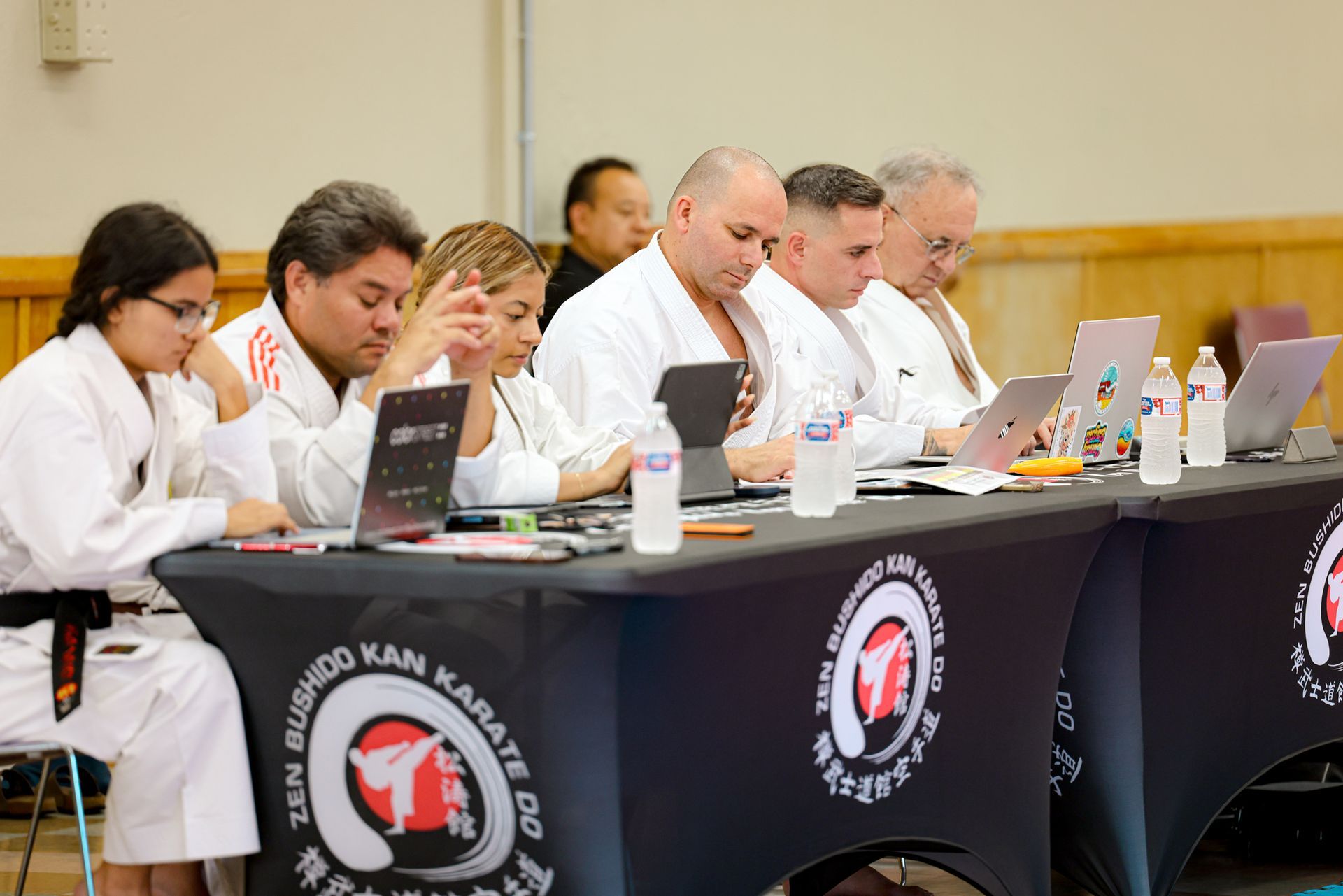 Judges in white uniforms review laptops and notes at a table during a martial arts competition.