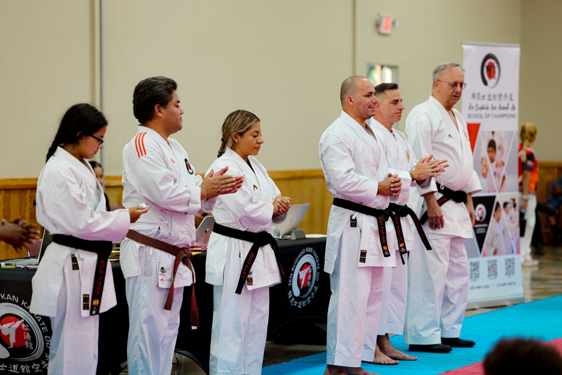 Martial arts instructors in white uniforms with black belts stand in a line at an indoor ceremony