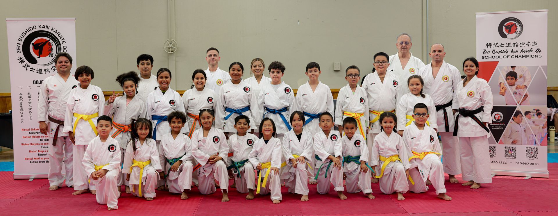 Martial arts group posing in white uniforms on a red mat, with banners in the background
