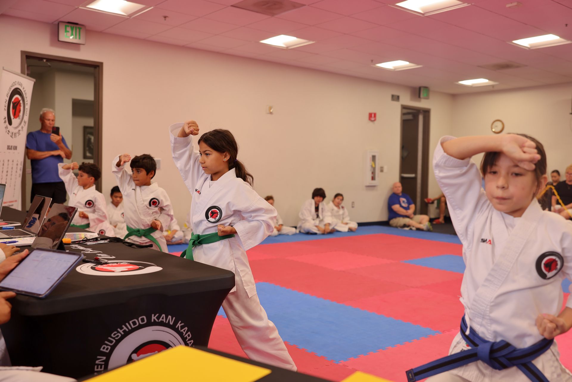 Karate class in a gym, children in white uniforms practicing on red and blue mats.