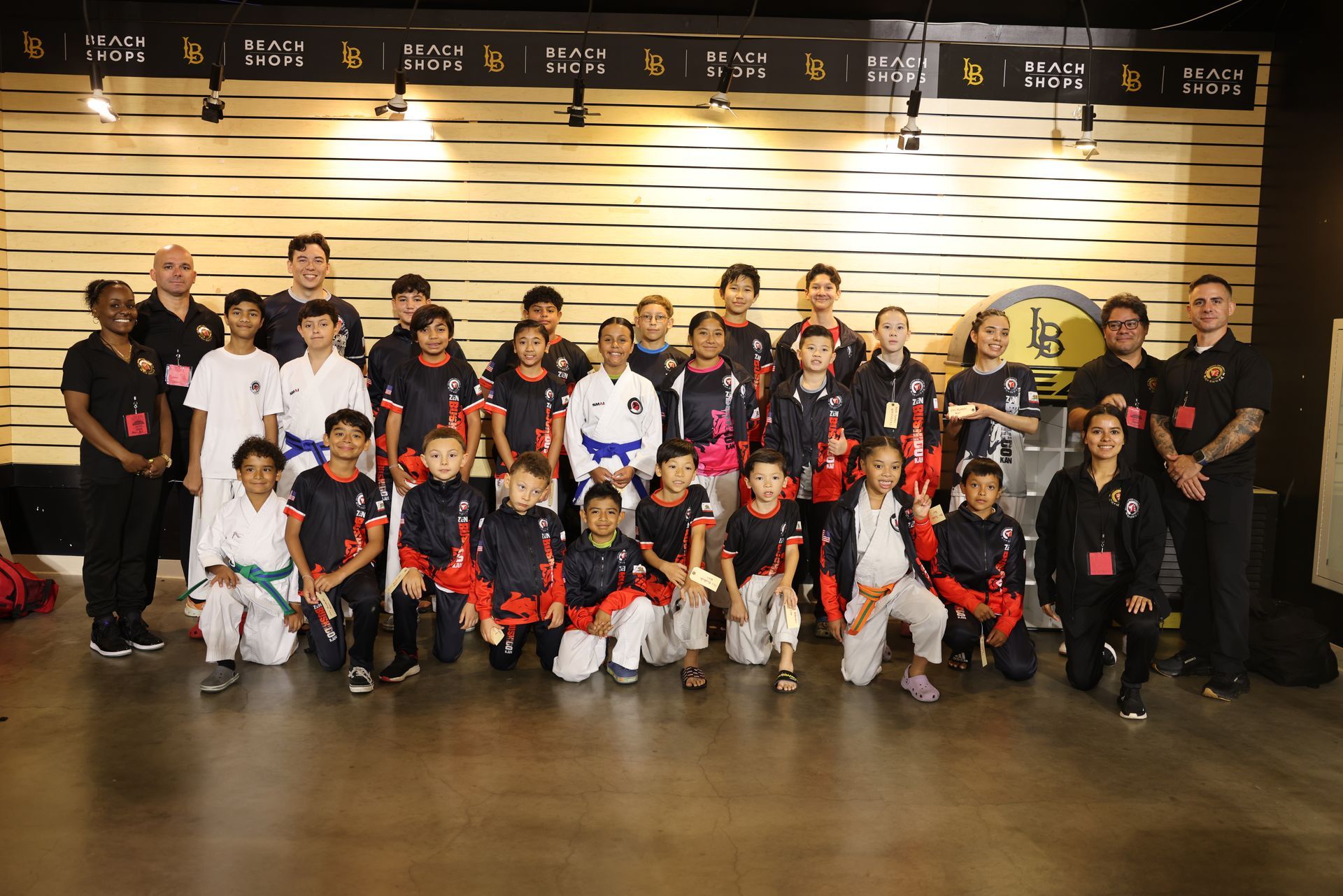 Group of martial arts students and instructors posing in uniforms indoors with trophies and banners behind them