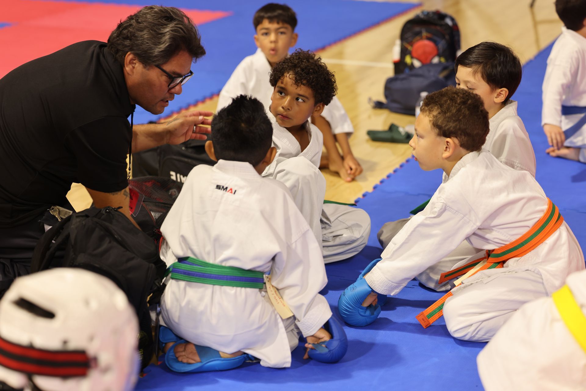 Martial arts instructor kneels with children in white gis on blue mats during a lesson