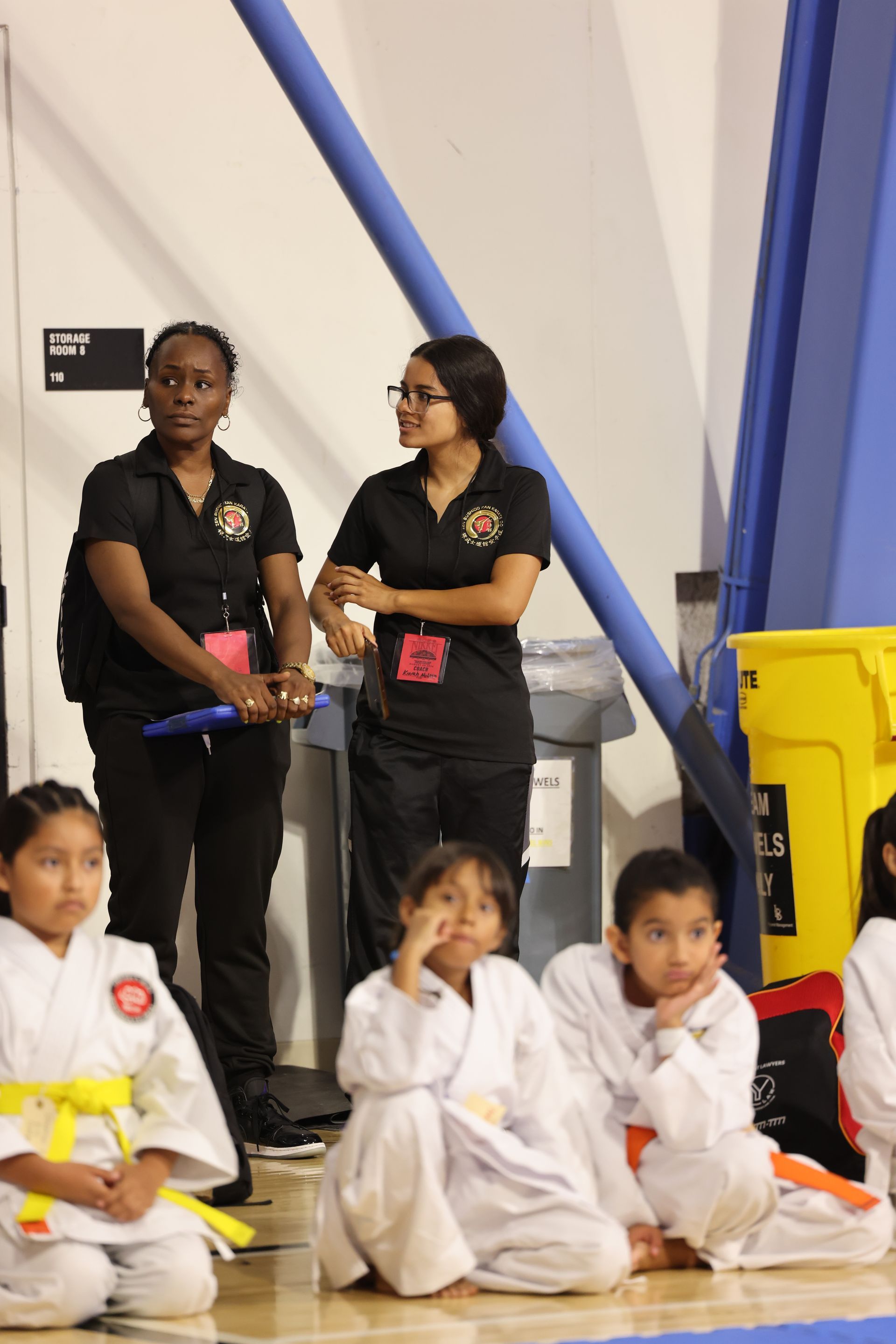 Children in white martial arts uniforms sit on a gym floor while two adults stand behind them.