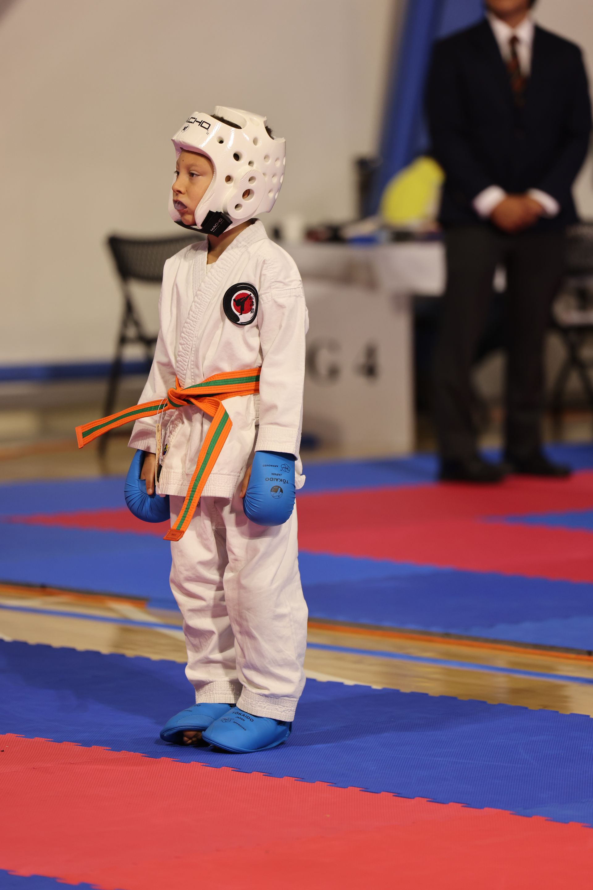Young karate competitor in white gi, blue gloves and helmet, standing on a red-and-blue mat during a match