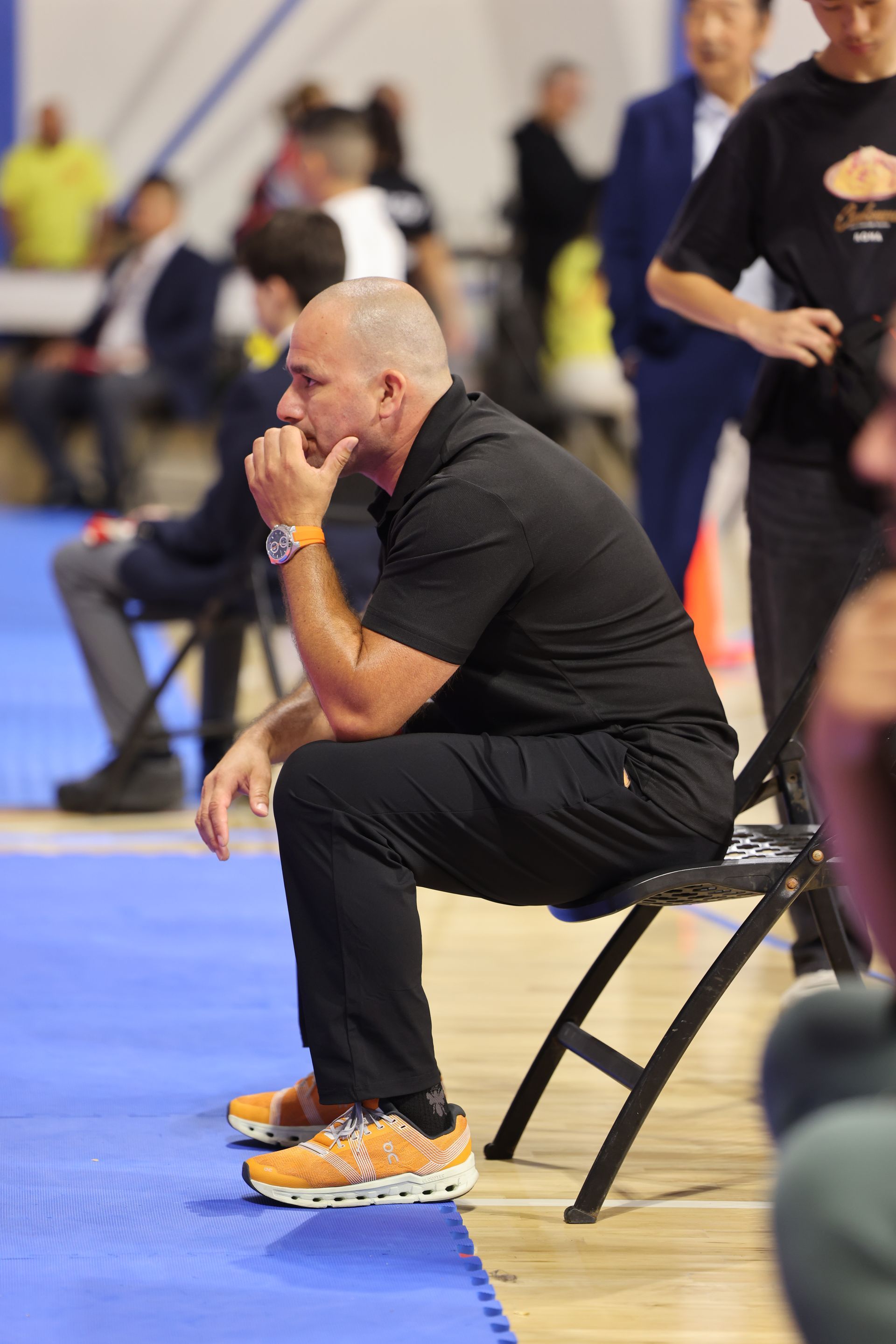 Man in black athletic clothes sits on a folding chair in a gym, watching an indoor sports event.