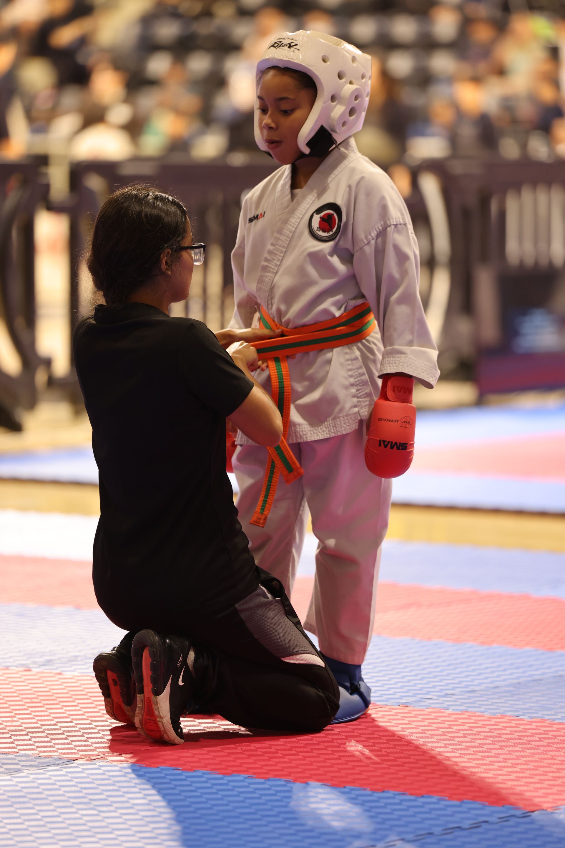 Martial arts match: child in white gi and red gloves stands while coach kneels beside them on a mat
