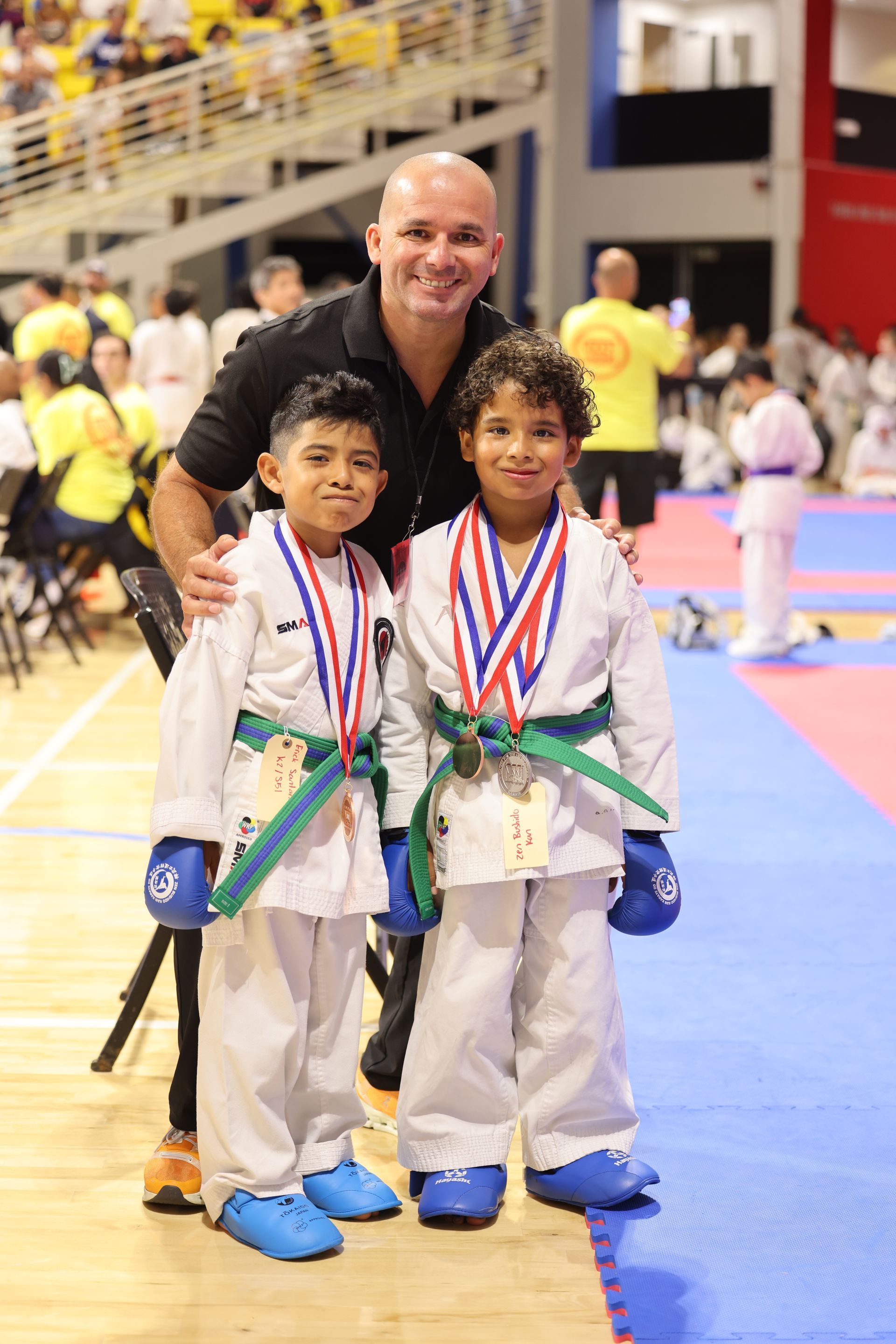 Two young karate competitors with medals pose beside their coach on a competition mat.