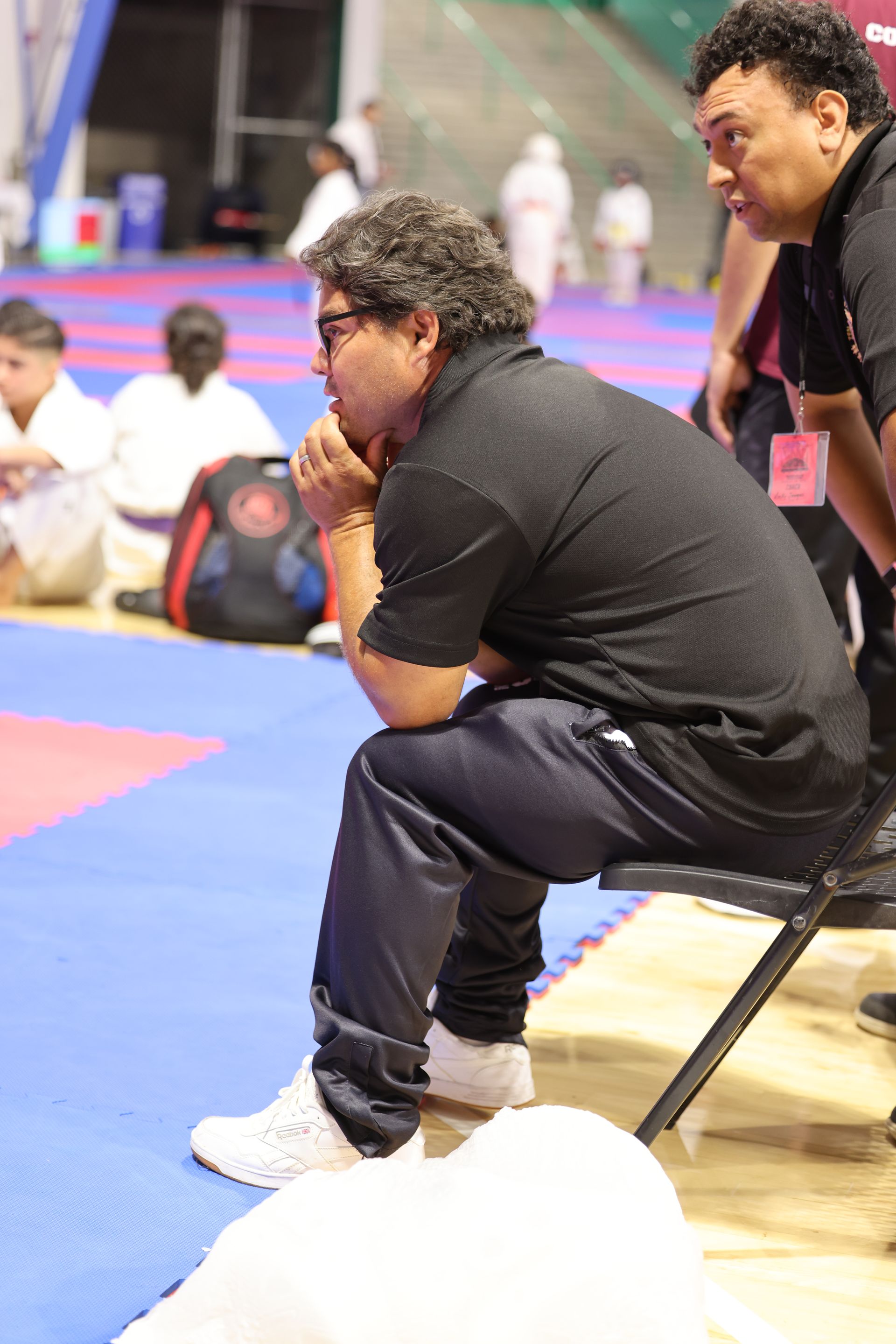 Man seated on a folding chair courtside at an indoor sports event, watching intently.