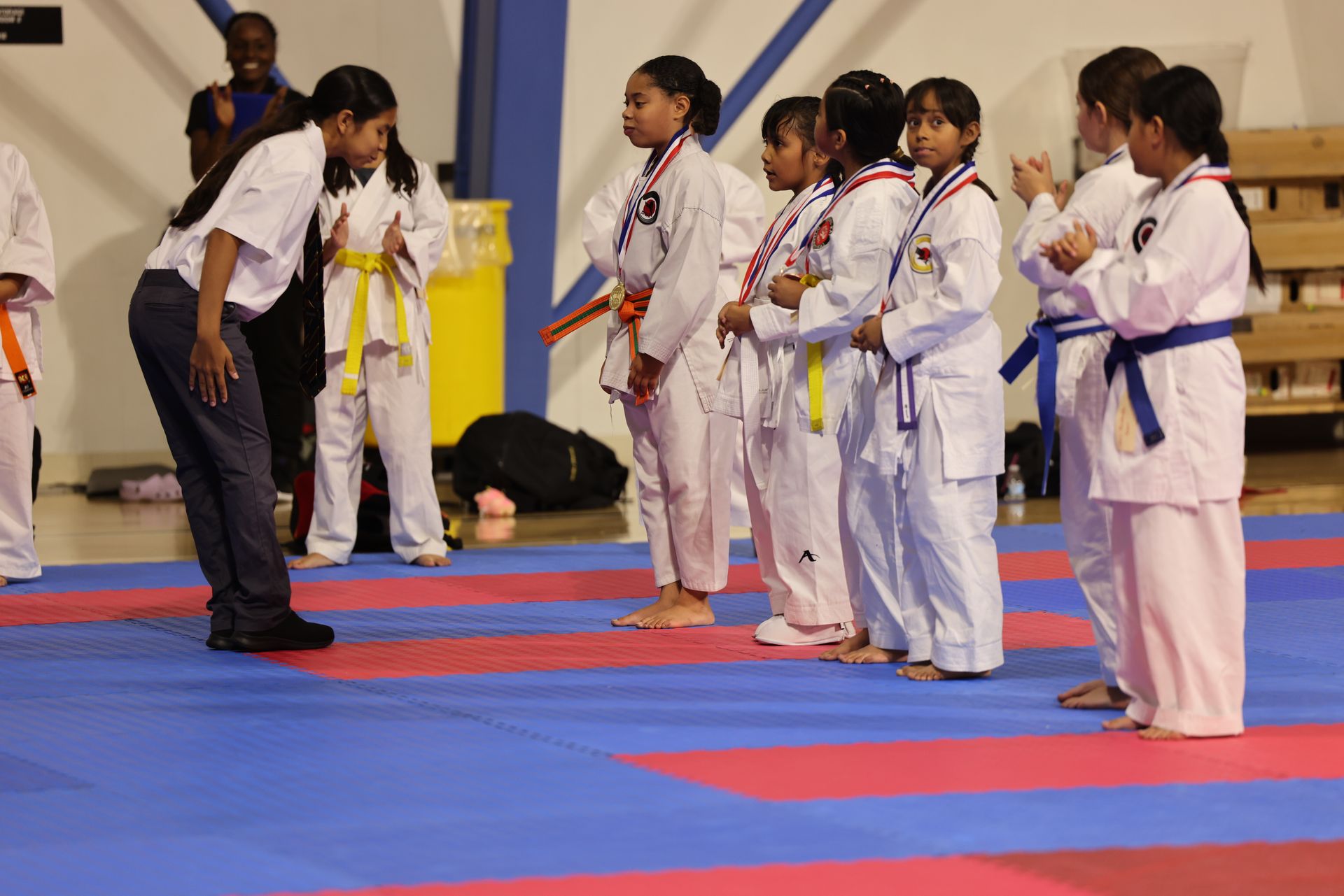 Martial arts instructor bows to a line of children in white uniforms with colored belts on blue and red mats
