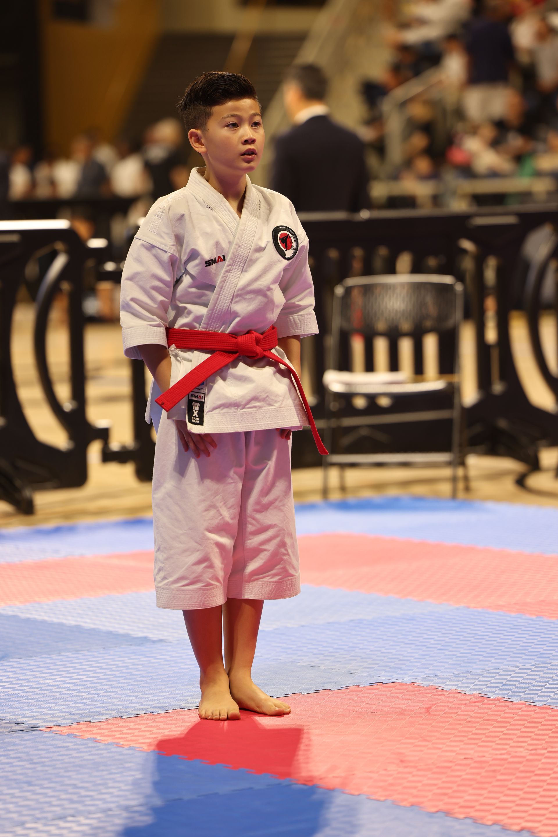 Karate competitor in white gi with red belt standing on a mat in an indoor arena