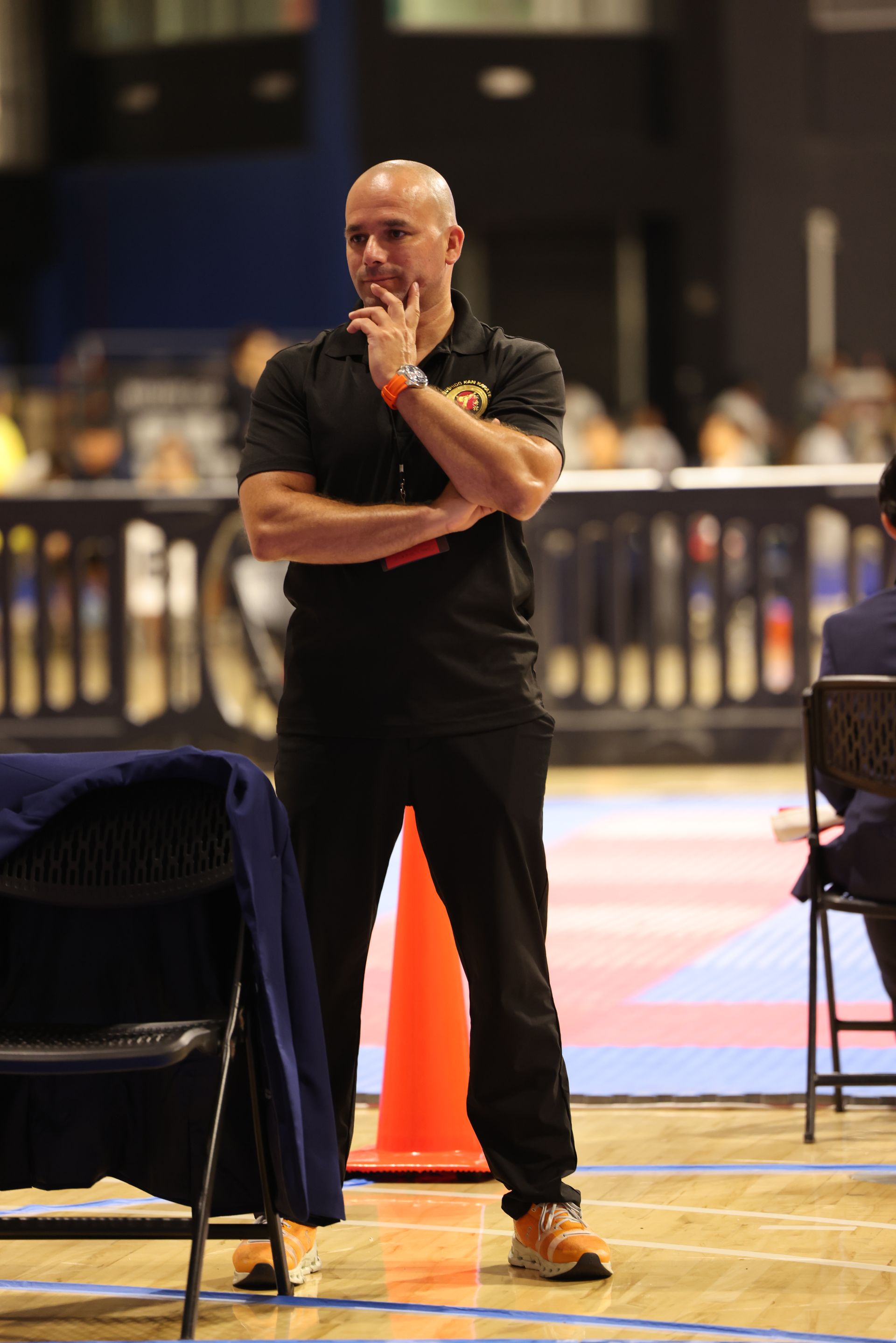 Man in black clothing stands on a gym floor with arms crossed, near an orange cone and folding chairs