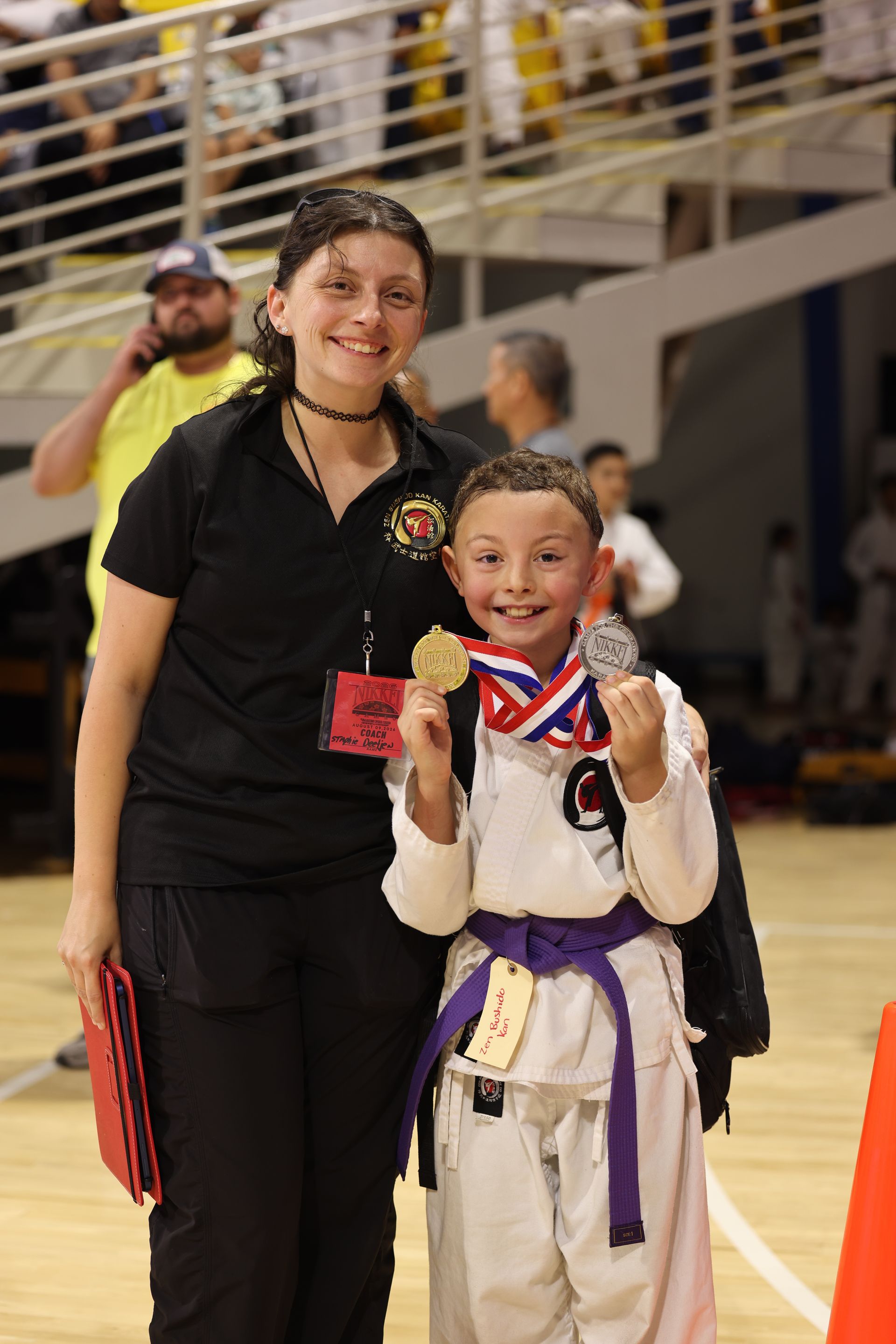 Two people in martial arts uniforms pose indoors, smiling and holding medals at a sports venue.