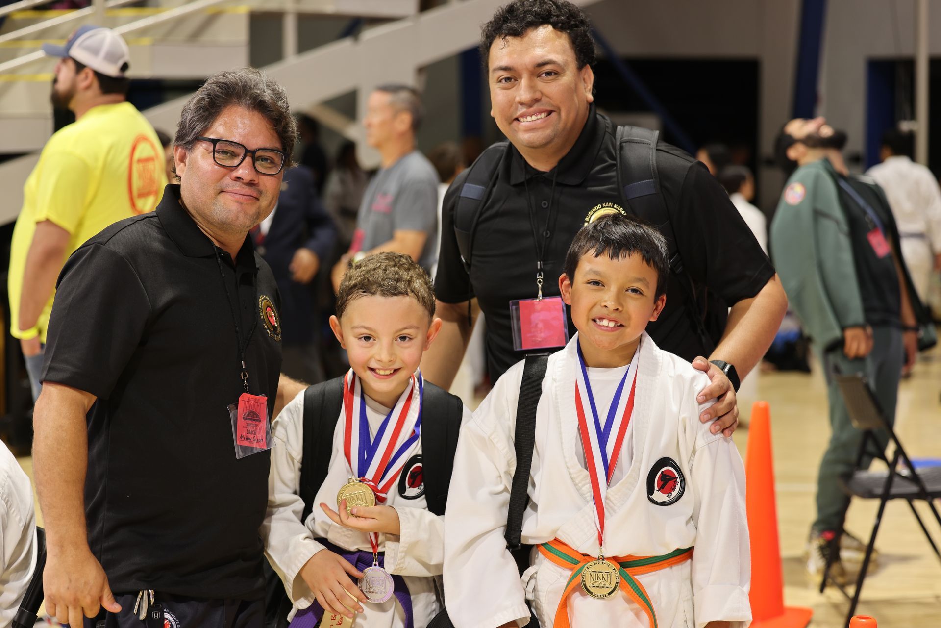 Two children in martial arts uniforms with medals, posing with two adults indoors at an event