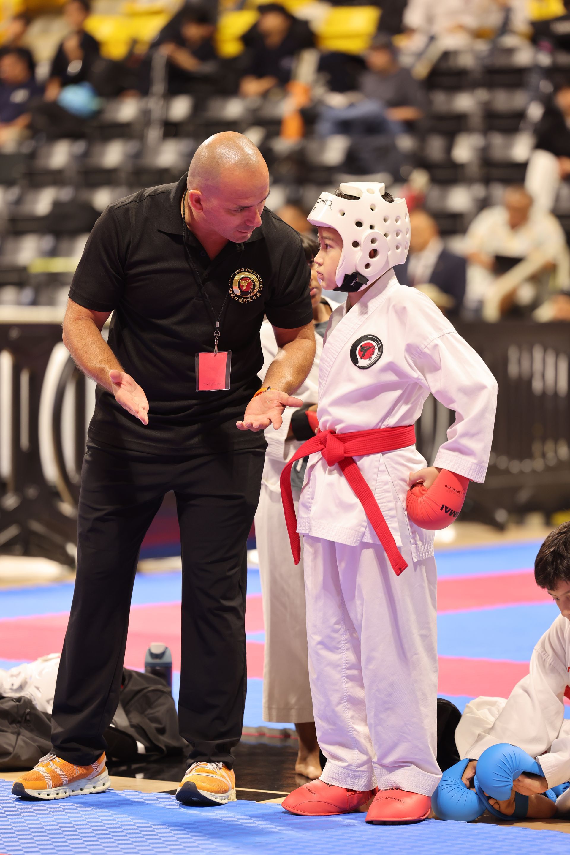 Martial arts coach speaking to a child in white gi and red belt on a competition mat