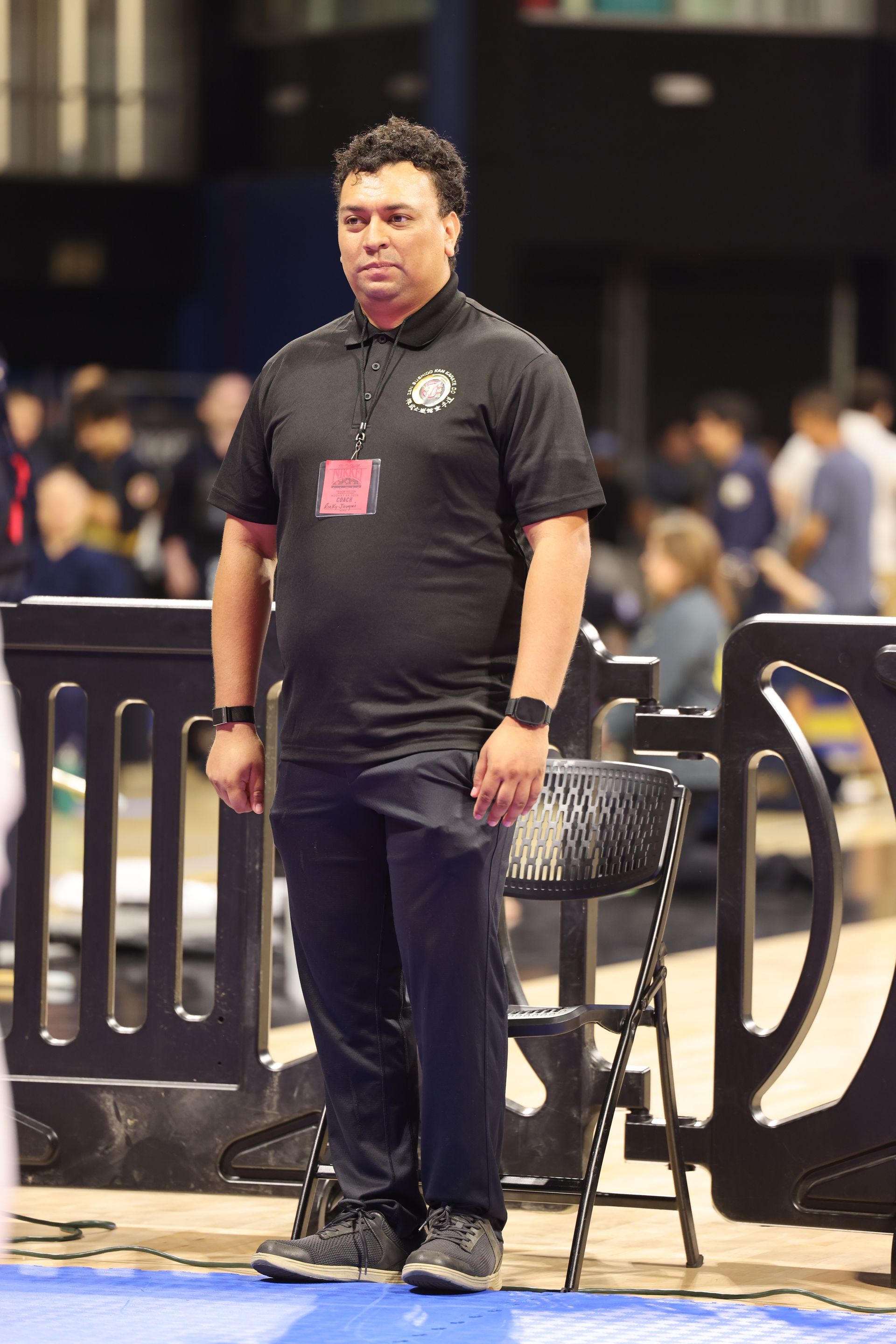 Man standing in a black polo and pants at an event, with chairs and people blurred in the background.