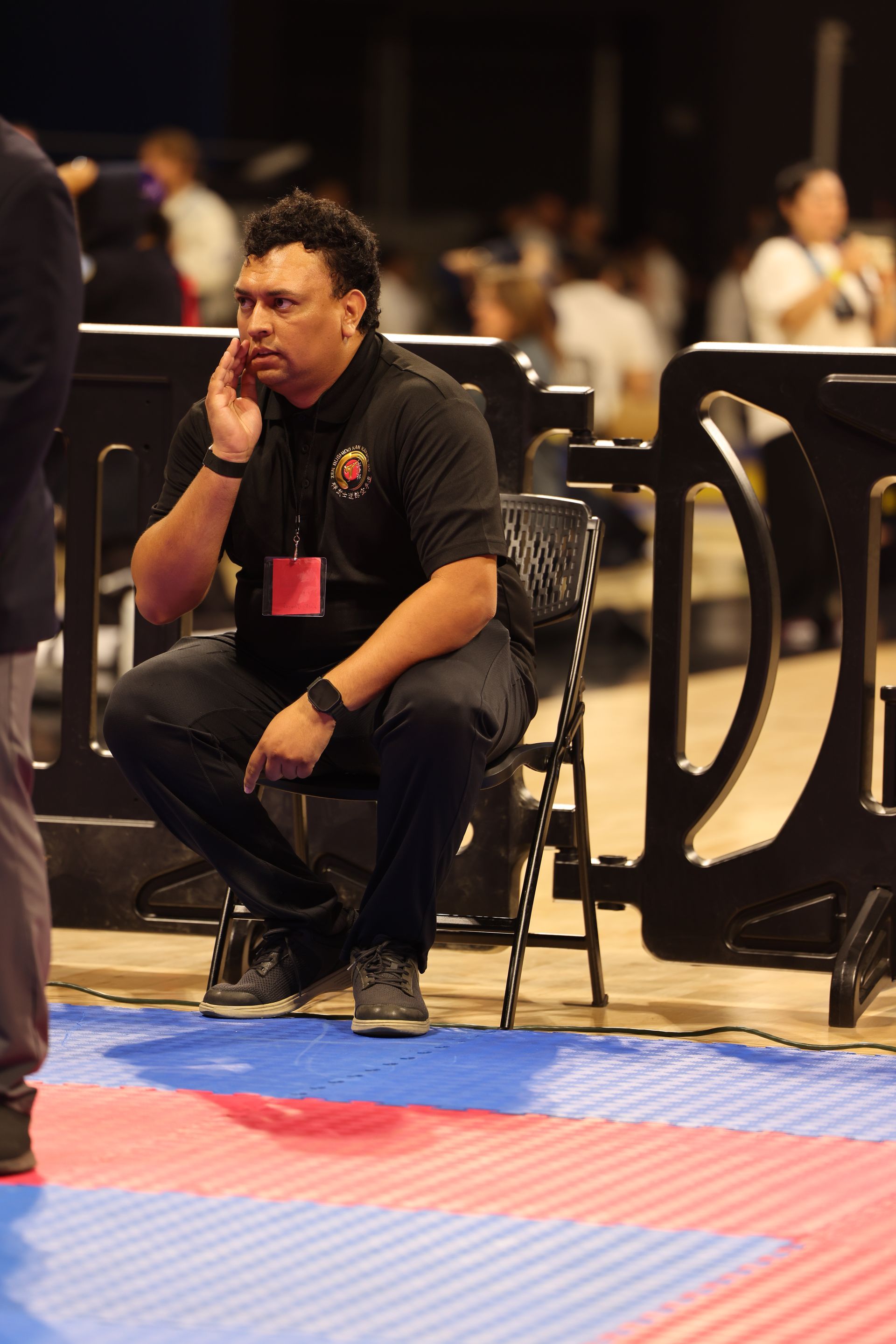 Person crouching beside a mat in an indoor sports venue, wearing black clothing and a red lanyard.
