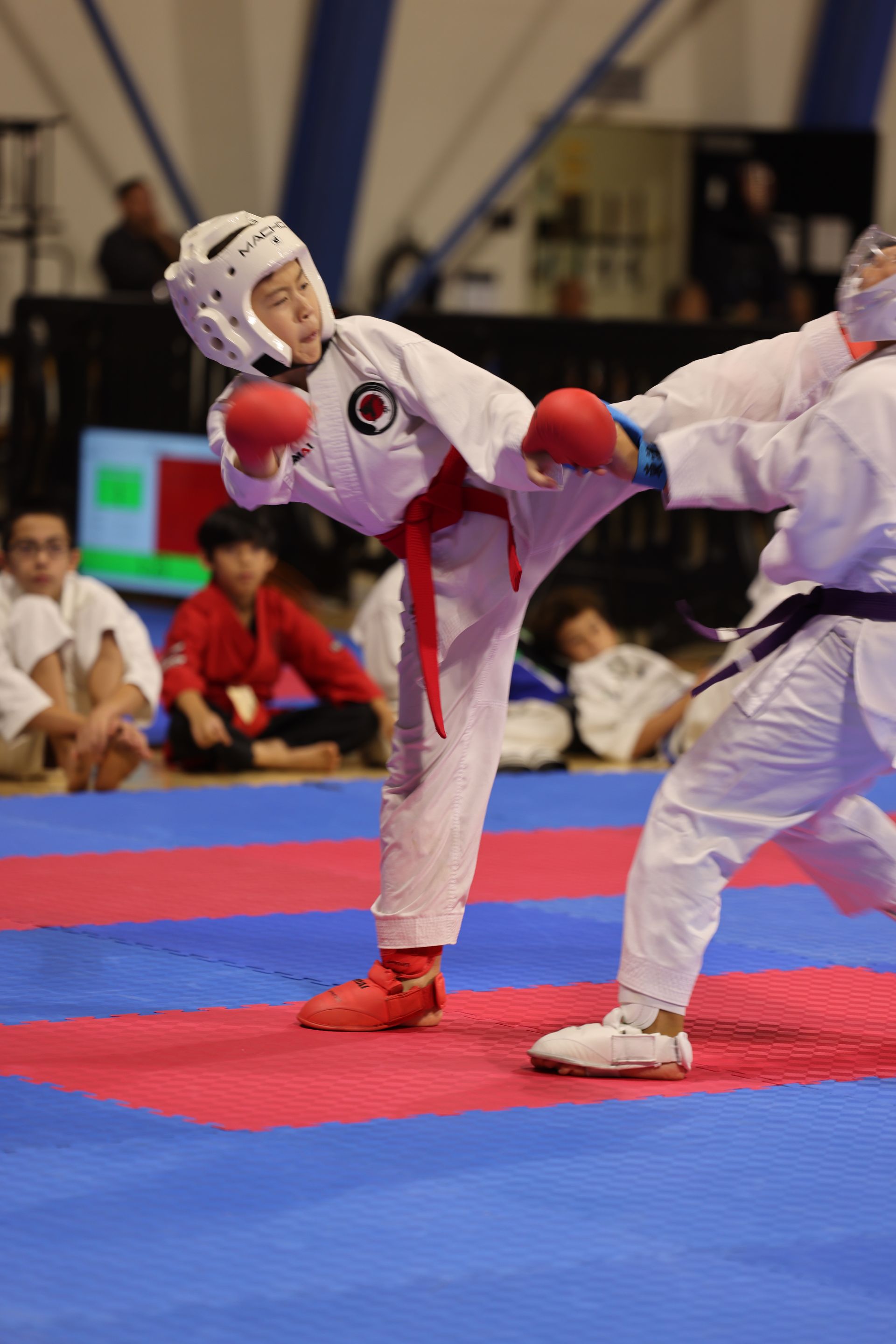 Two martial artists sparring; one in white gear kicks the other on a red-and-blue mat indoors.