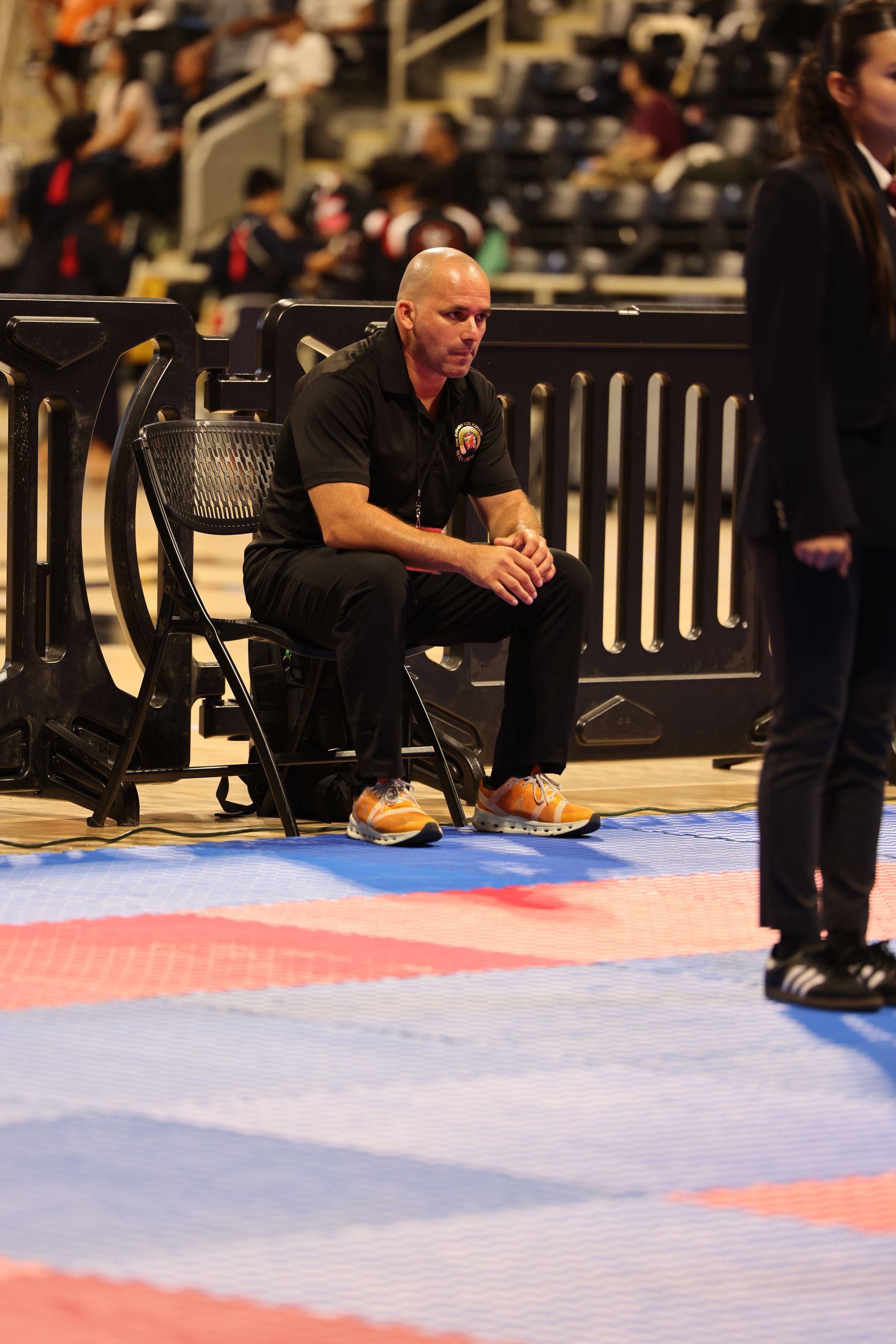 Man sitting courtside near a black barrier at an indoor sports event, watching the action.