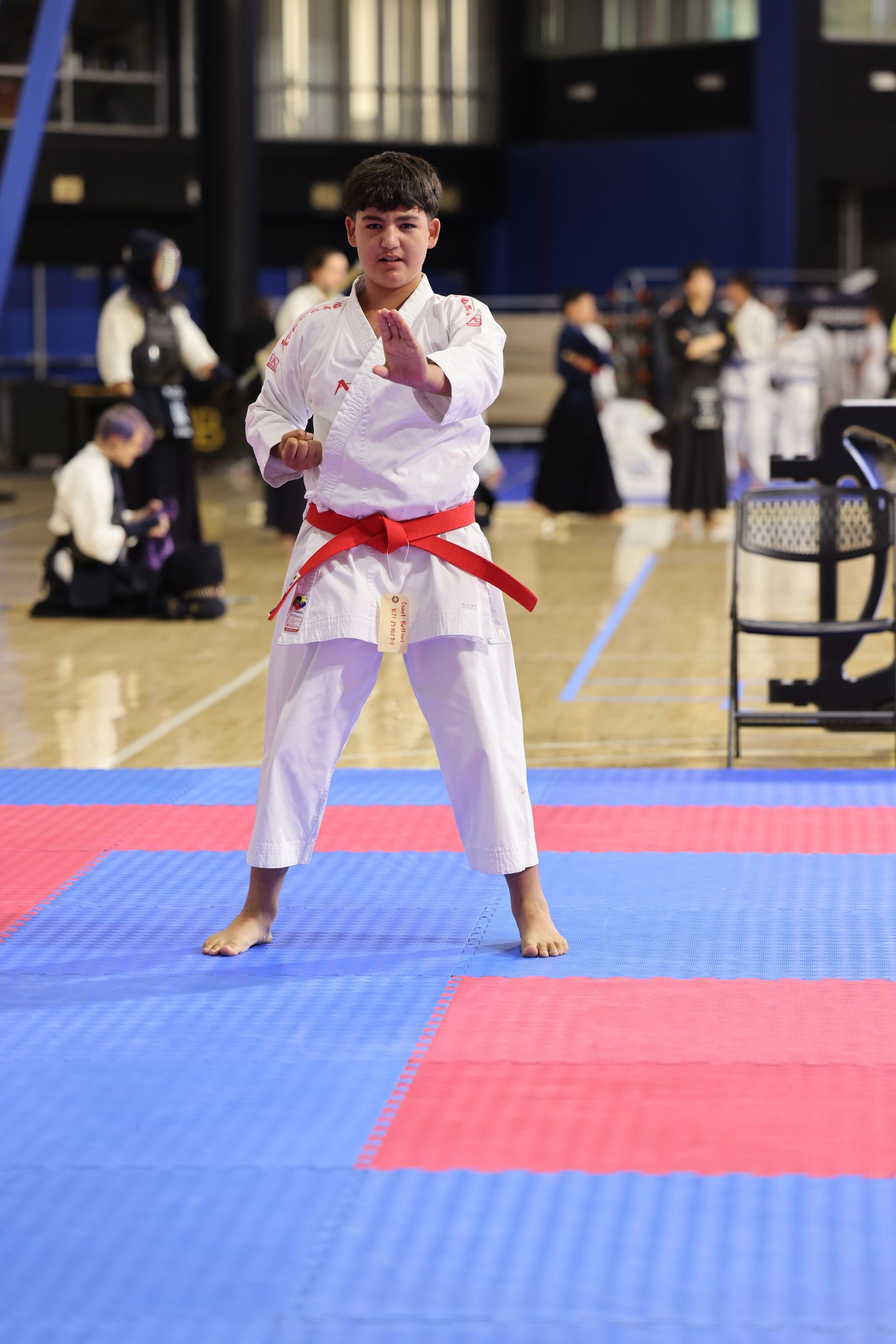Karate competitor in white gi with red belt stands on blue and red mats in an indoor gym.
