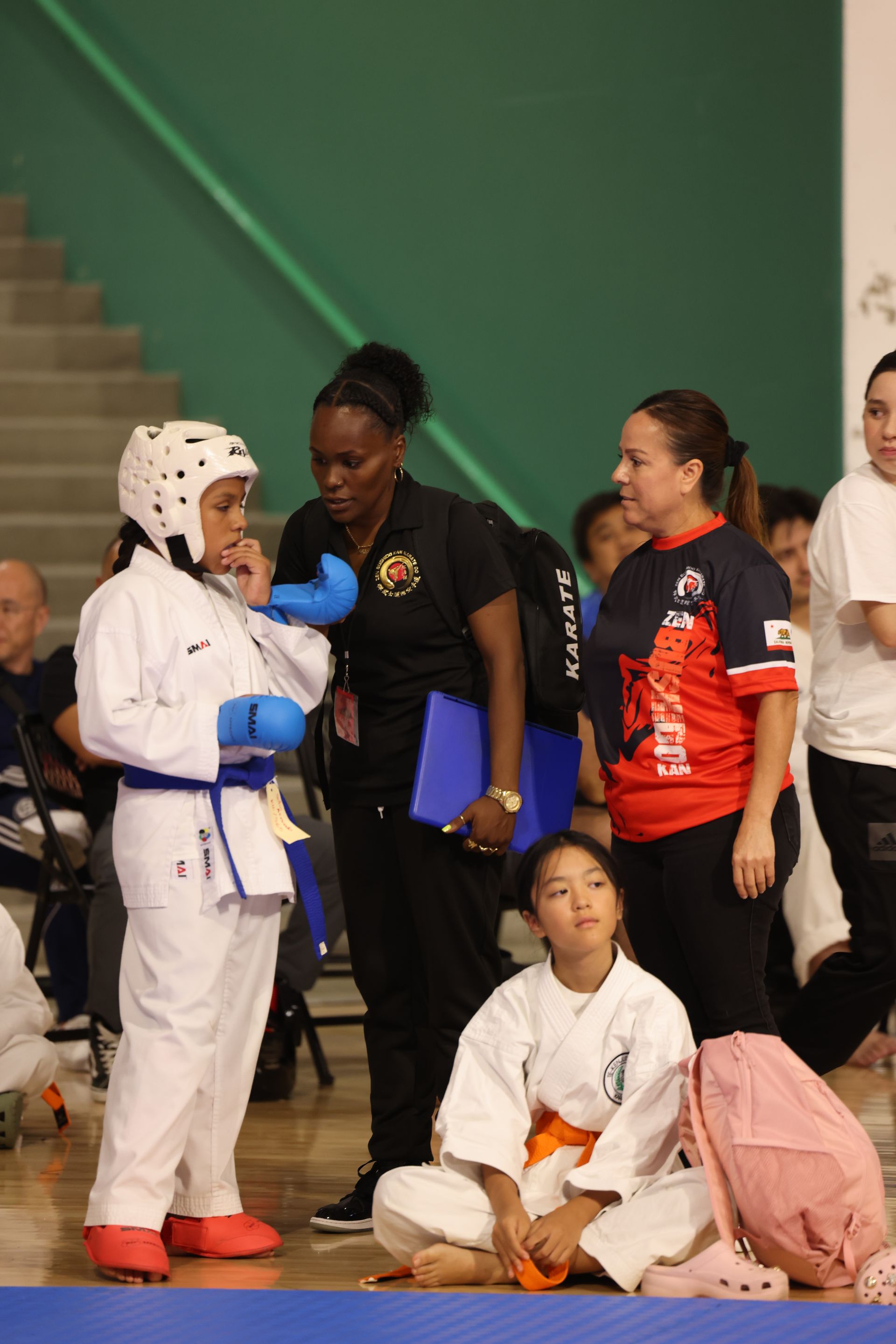 Martial arts competitors and coach in a gym, with one person kneeling in a white uniform and blue belt.