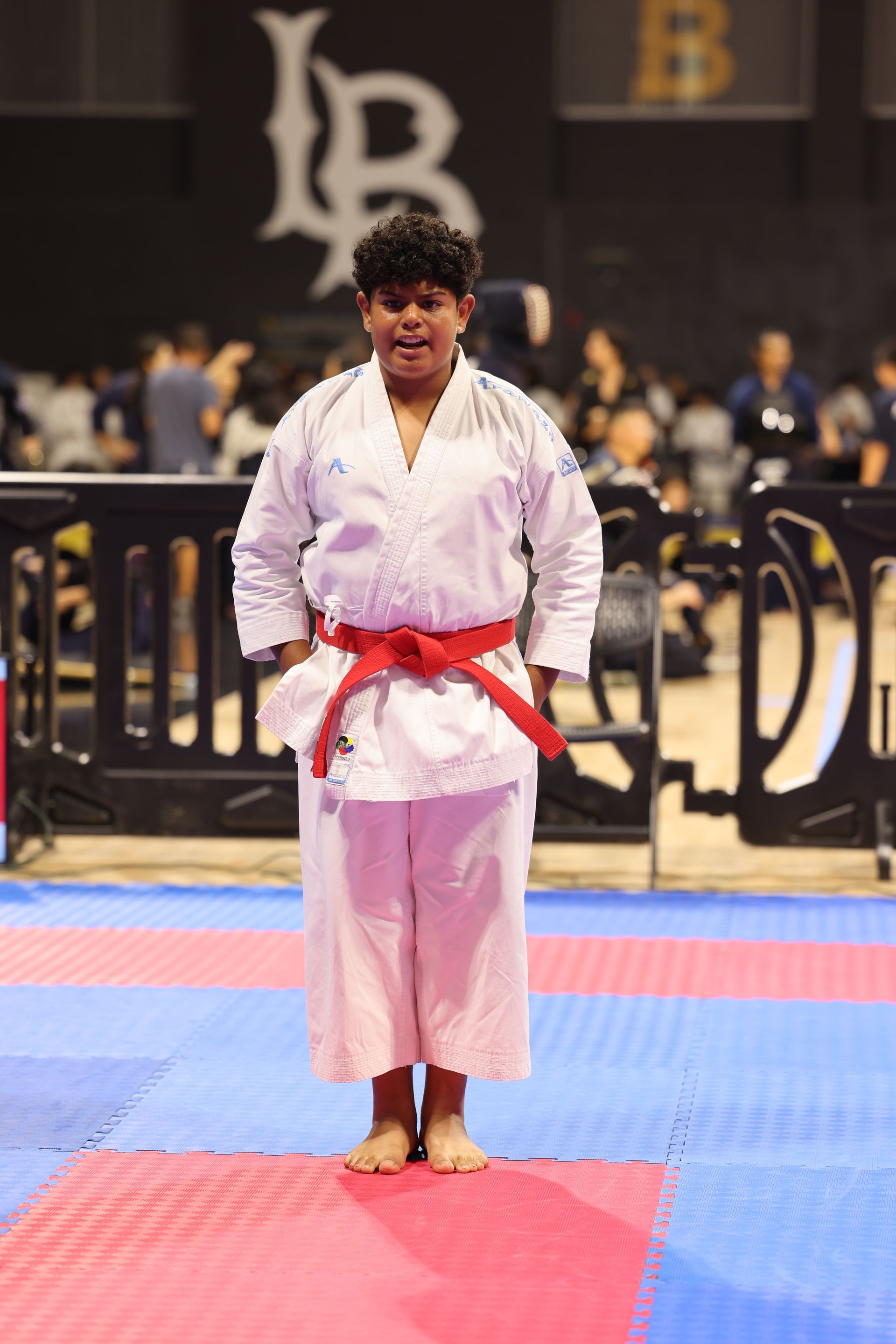 Martial artist in a white gi with red belt stands on a competition mat in an indoor arena