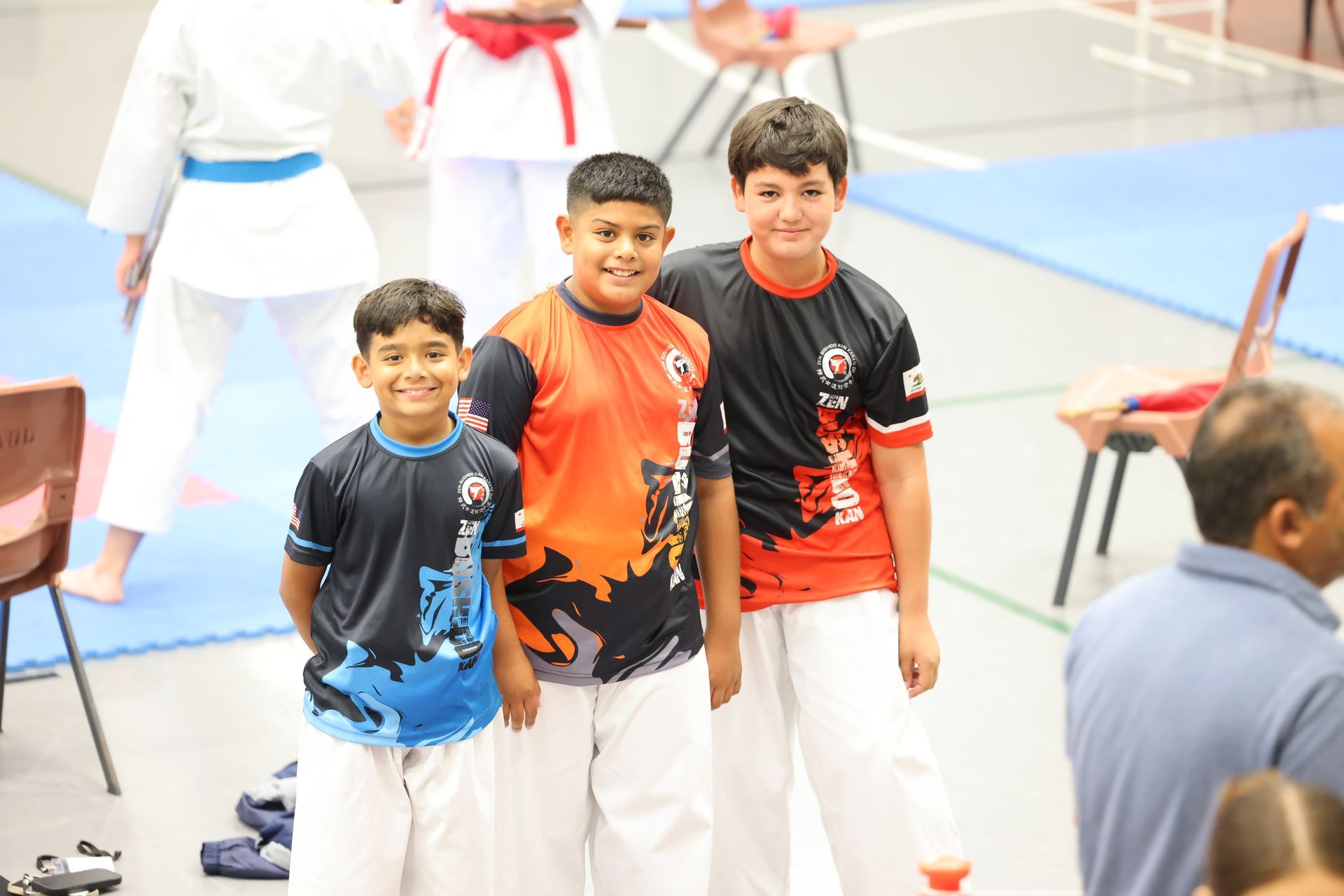 Three young boys in karate uniforms smiling at a tournament. Two in team shirts, one in blue.