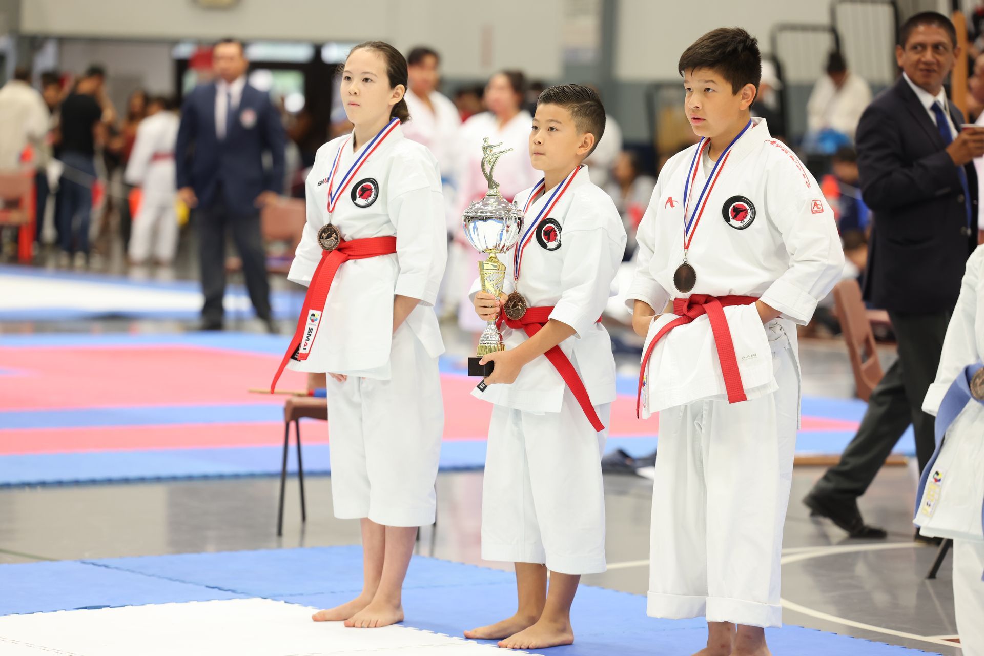 Karate kids on a podium with medals and trophy, wearing red belts and white gis, in a competition hall.