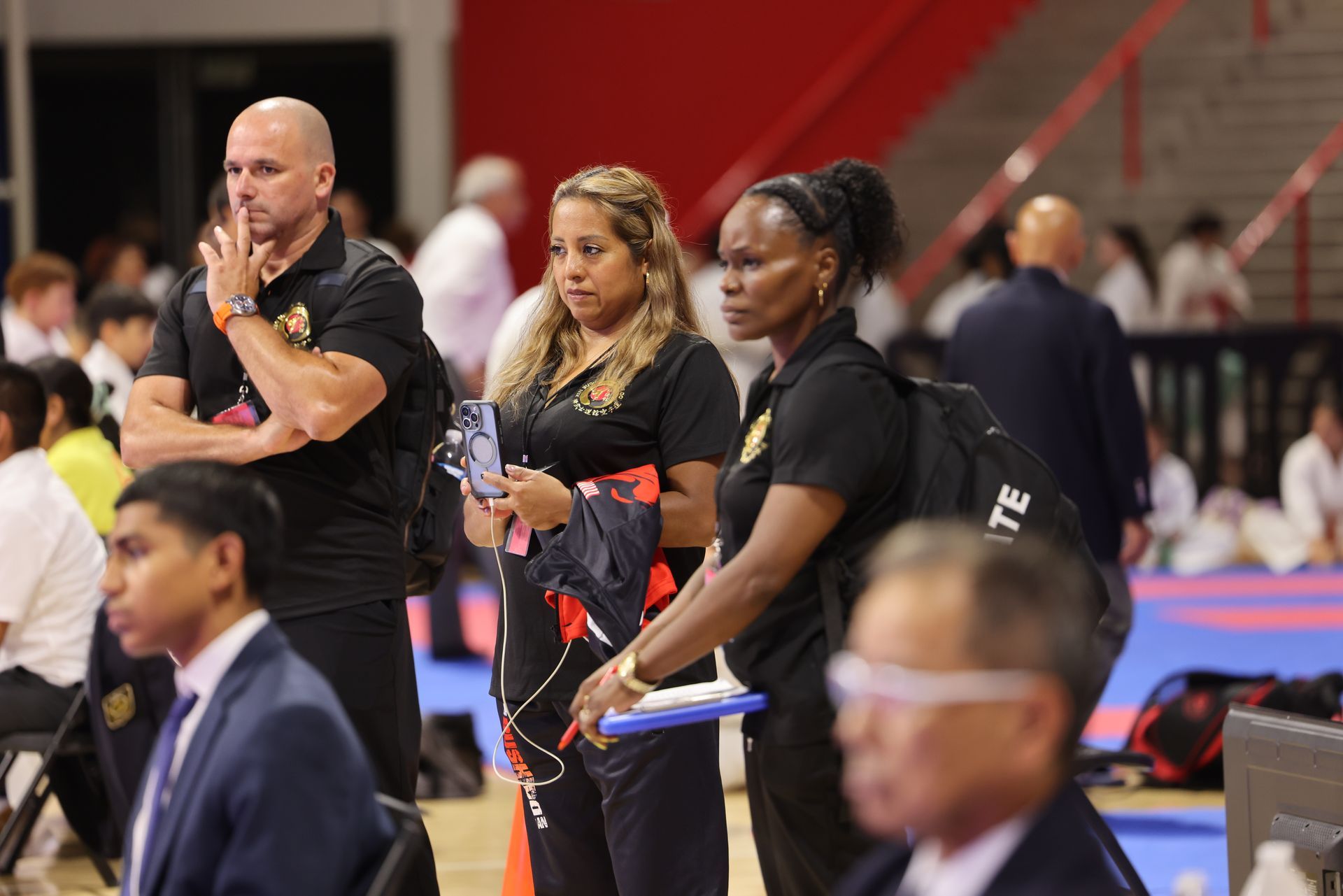 People standing at the edge of a martial arts mat, watching an event in a gymnasium