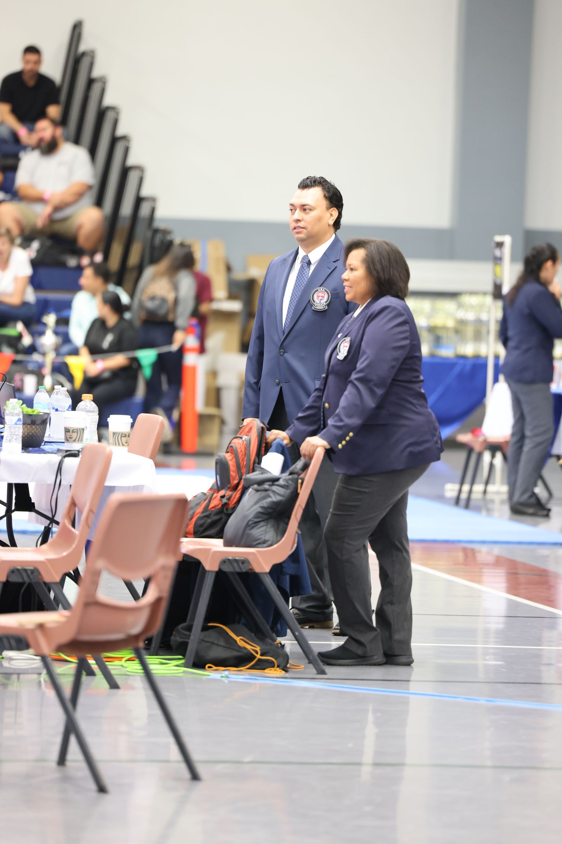 Man and woman in blazers at event, standing near chairs. Indoor venue with audience.