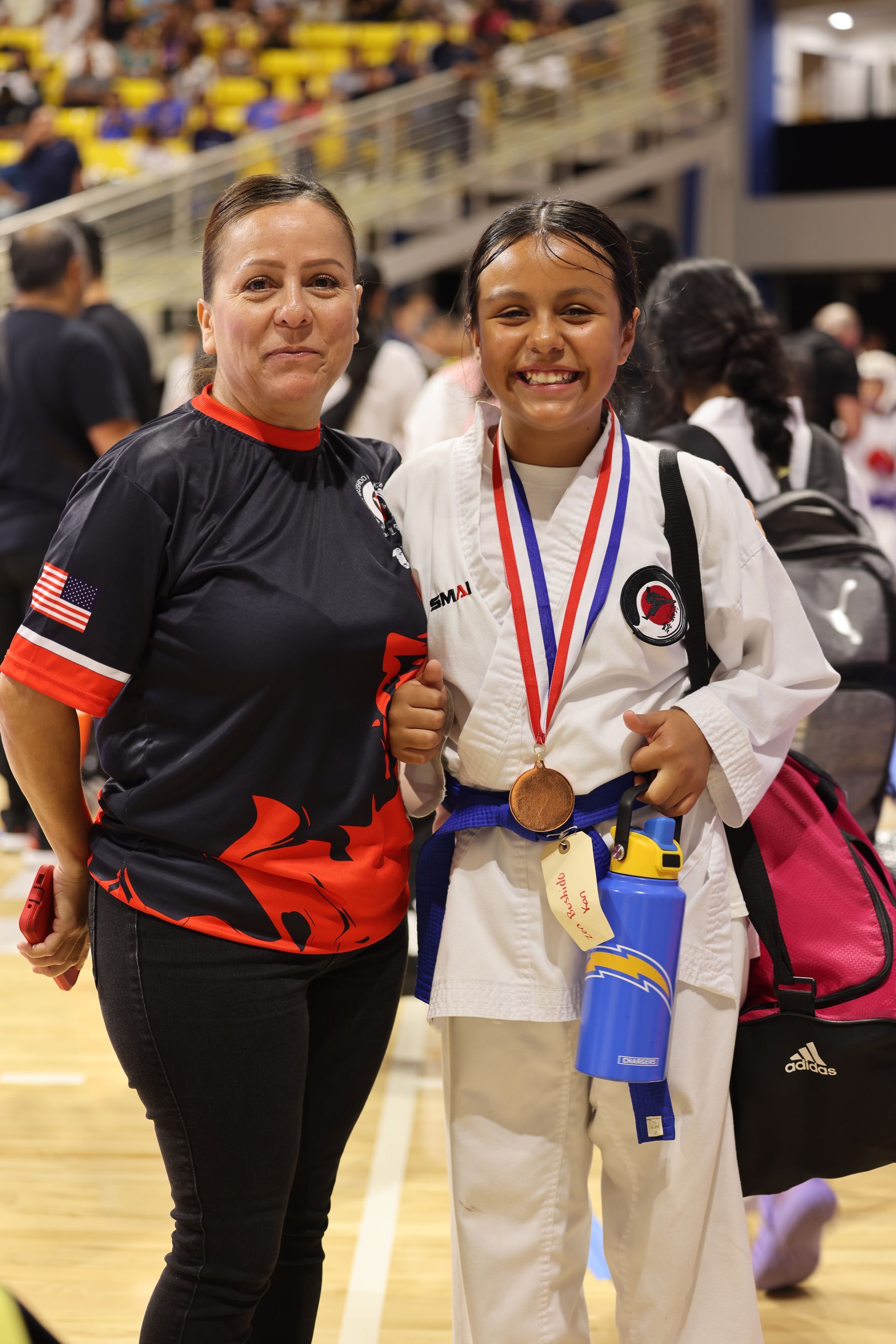 Two martial artists pose with medals at an indoor competition, one in a white gi and one in a black shirt.