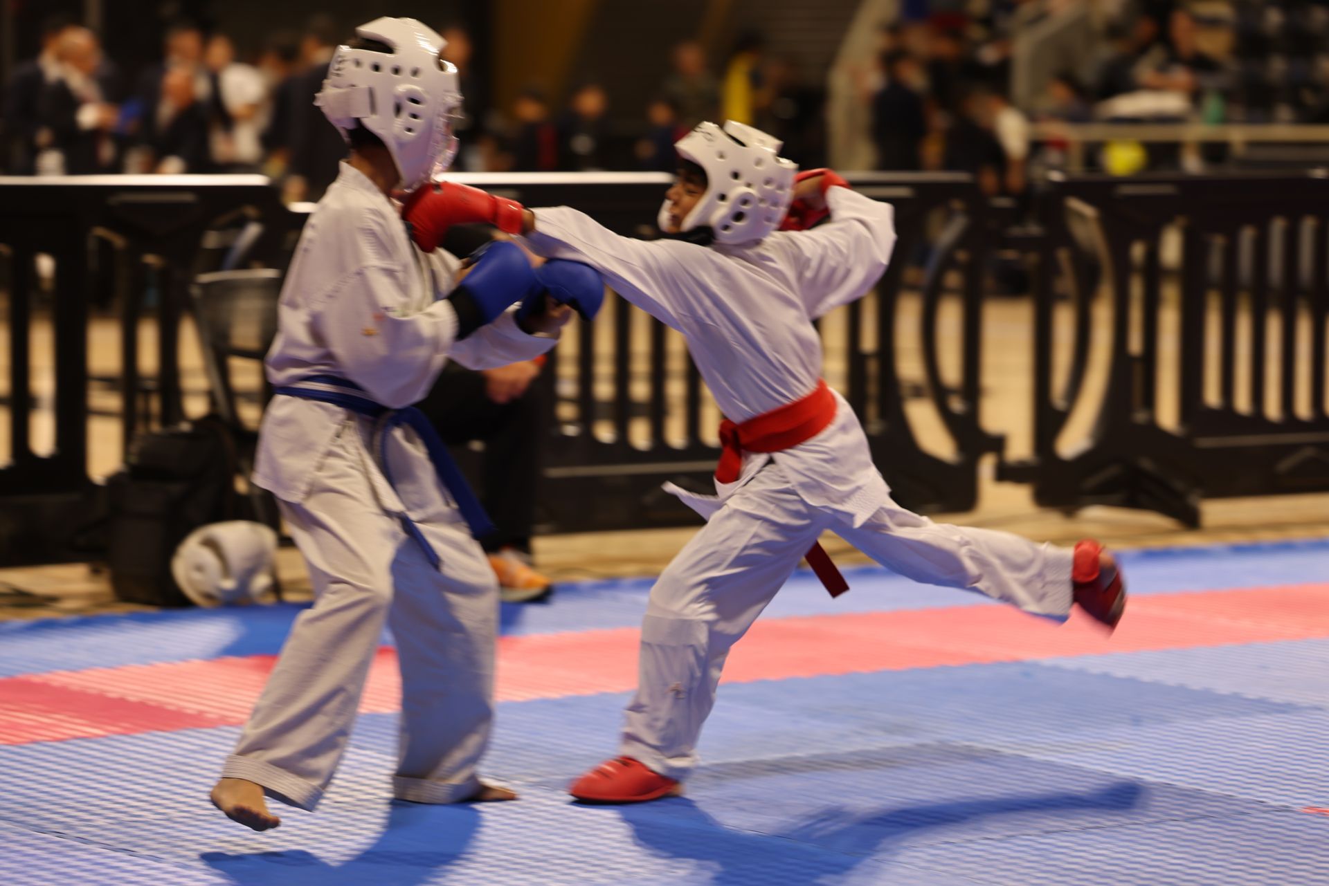 Two children sparring in karate gear on a blue mat, one kicking and the other blocking.