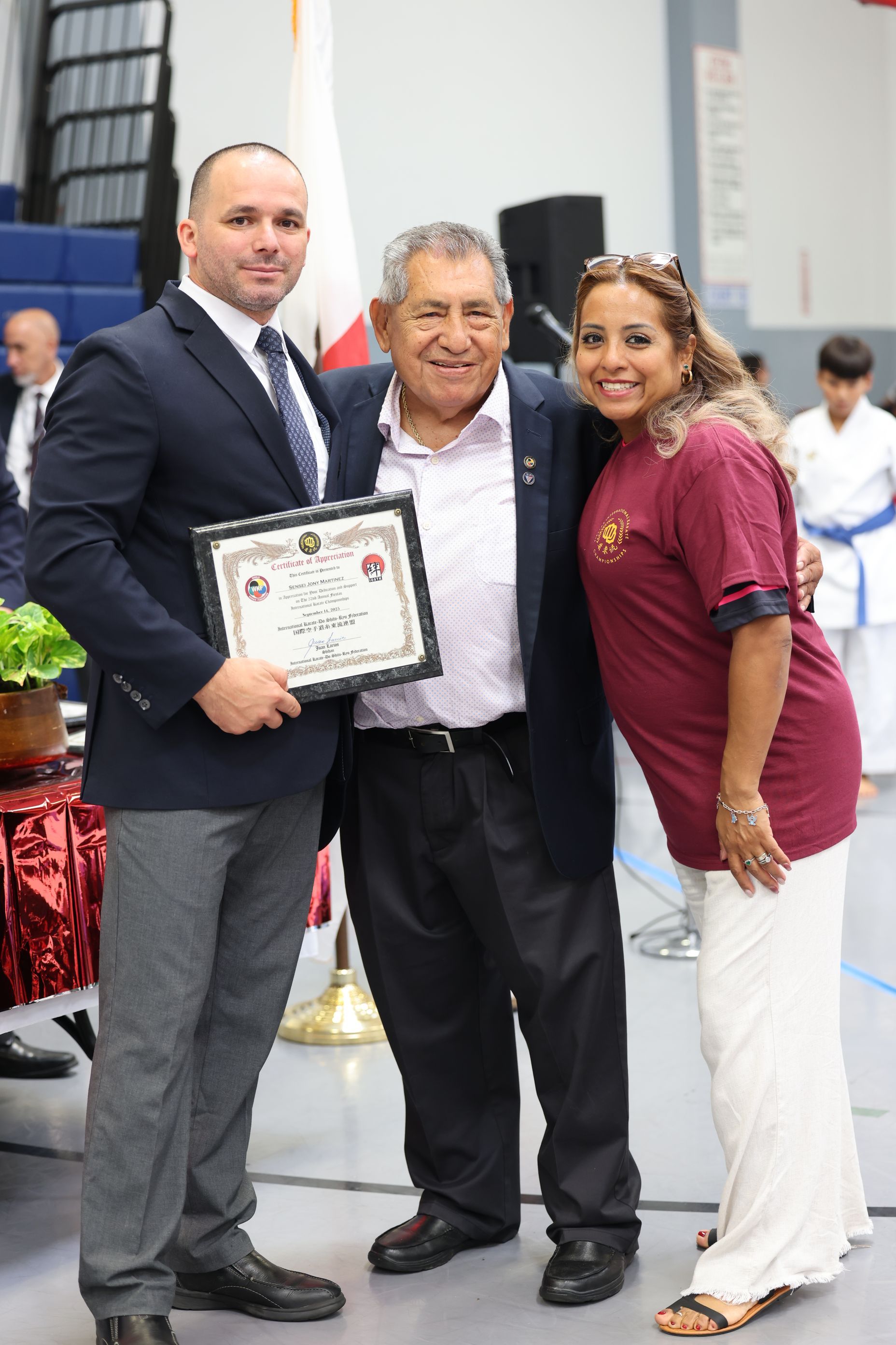 A man in a suit holds a framed certificate, smiling. A man in a jacket smiles between them. Woman hugs them both.