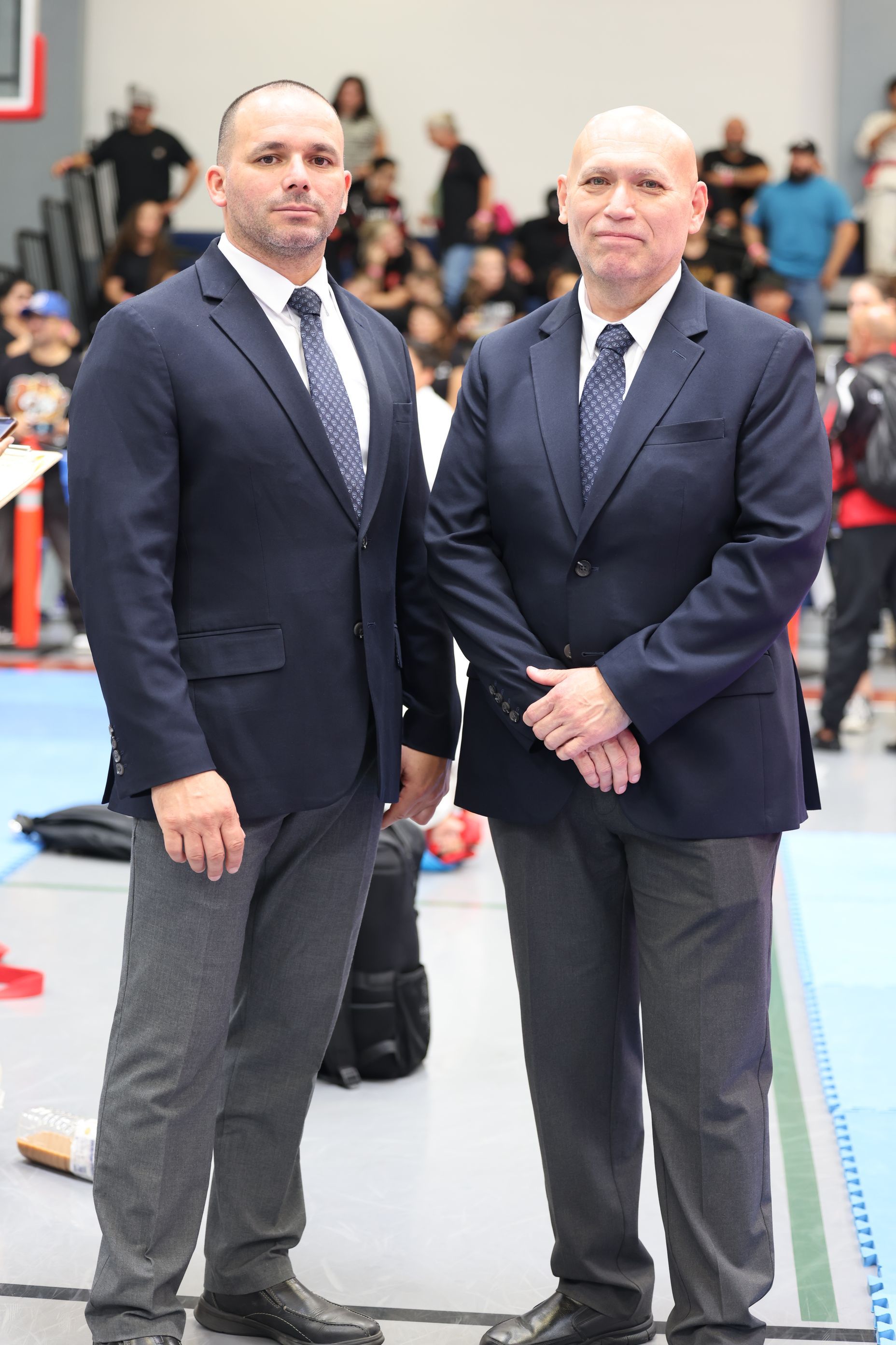 Two men in suits stand indoors, likely at an event. They appear serious.