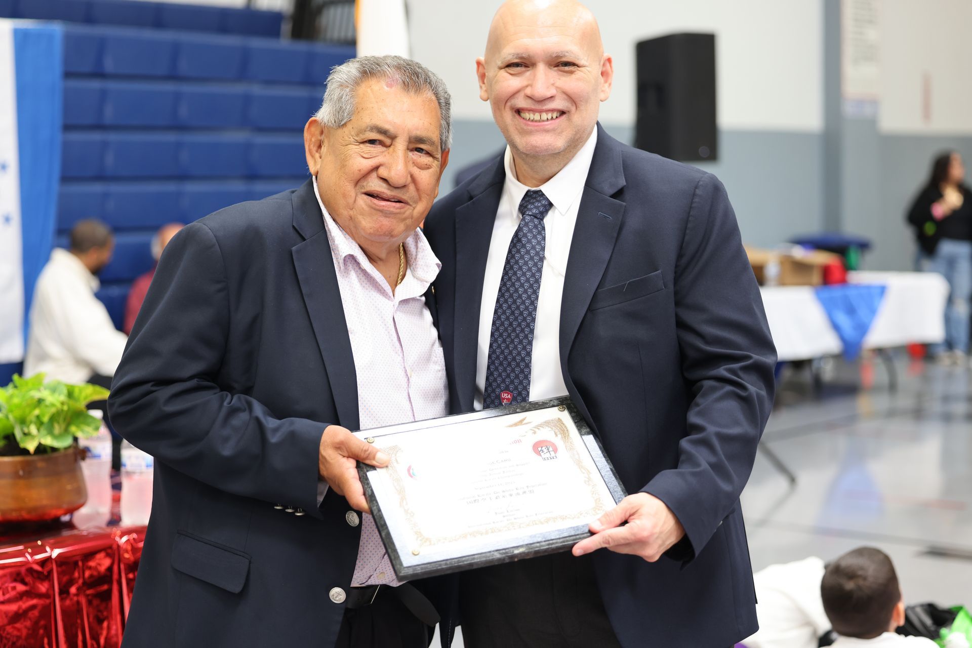 Two men smiling, holding a certificate, in a gymnasium. Older man in blazer, younger man in suit.