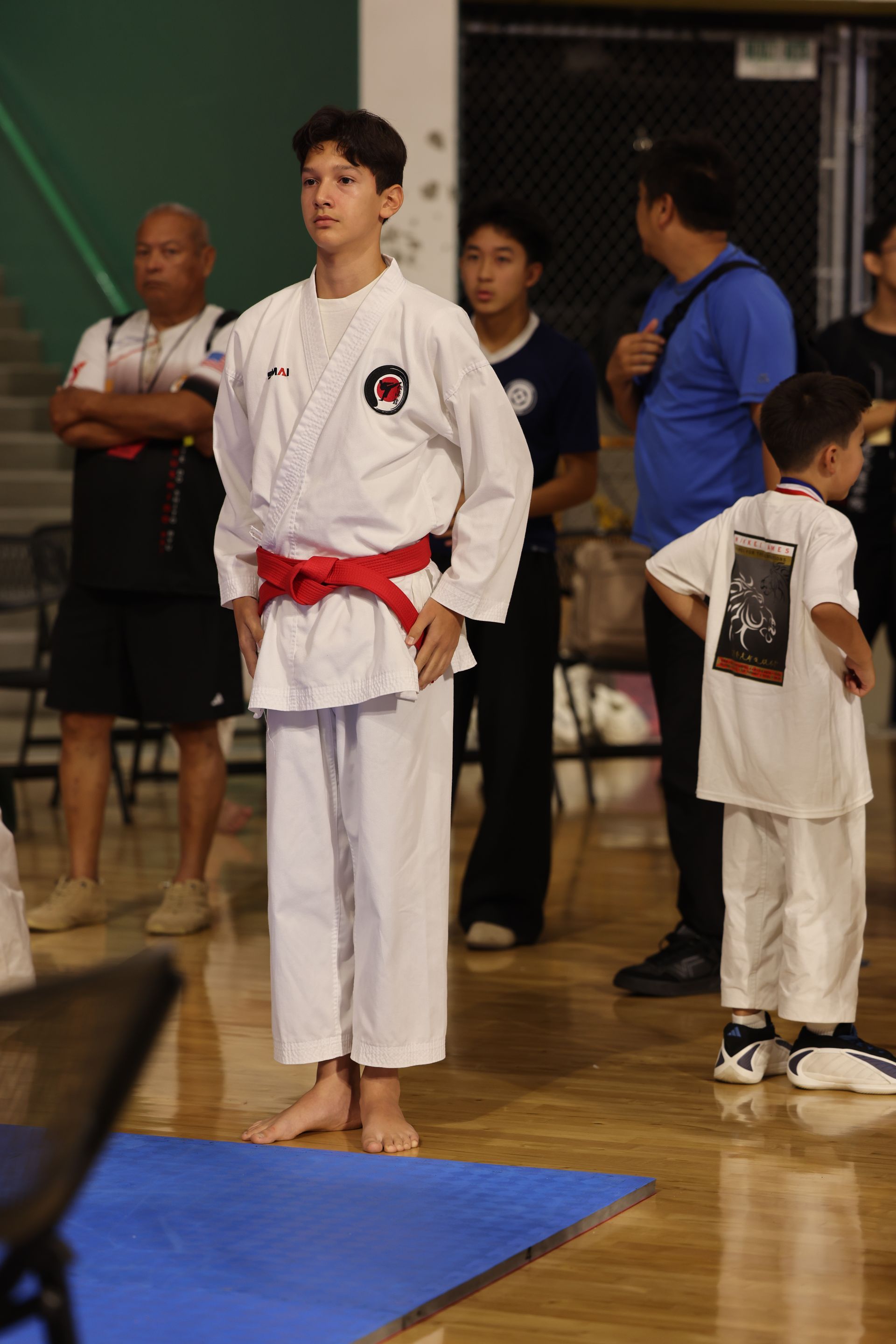 Martial arts student in white gi with red belt stands on mat at an indoor gym competition