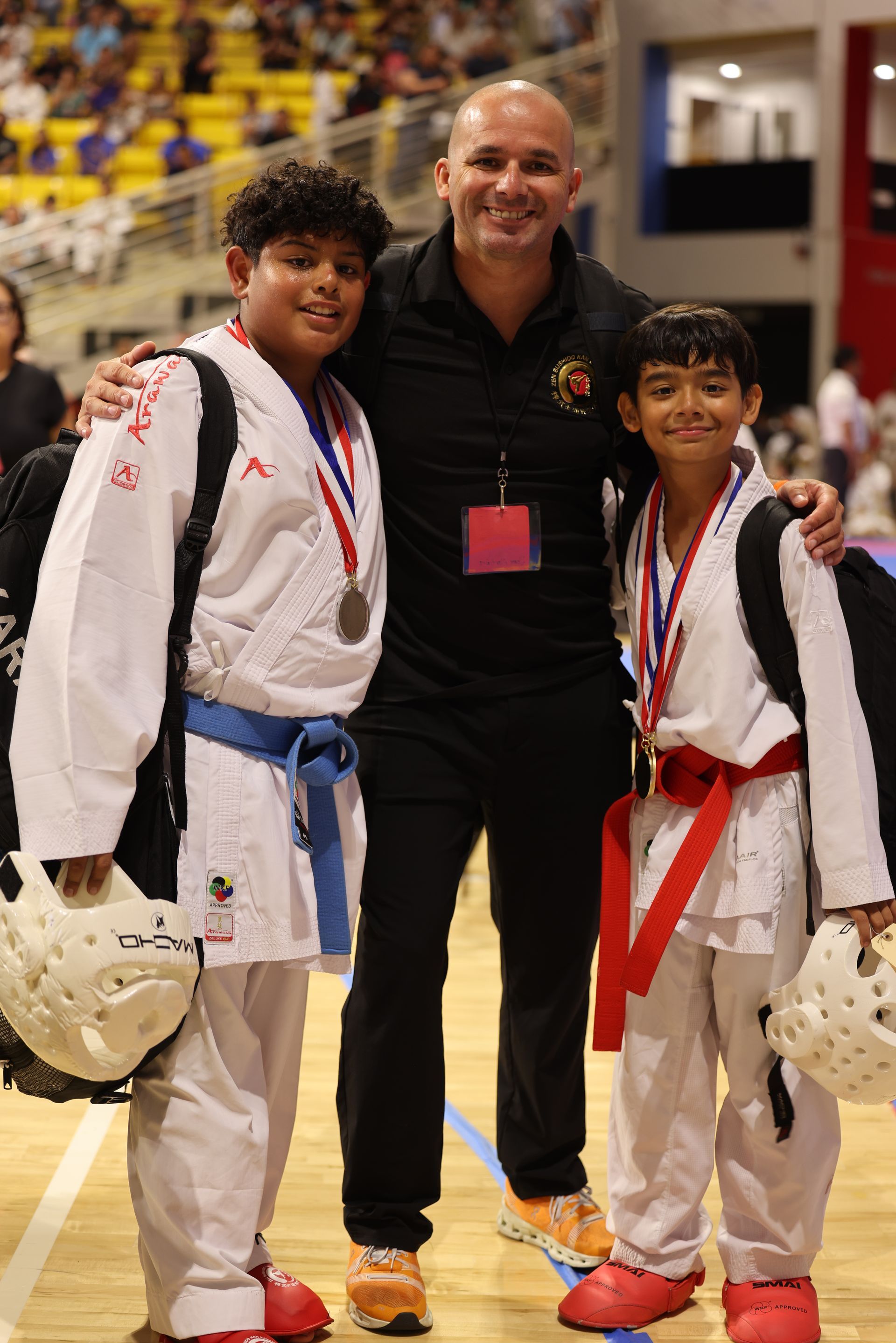 Three people posing in karate uniforms indoors, smiling and wearing medals and helmets
