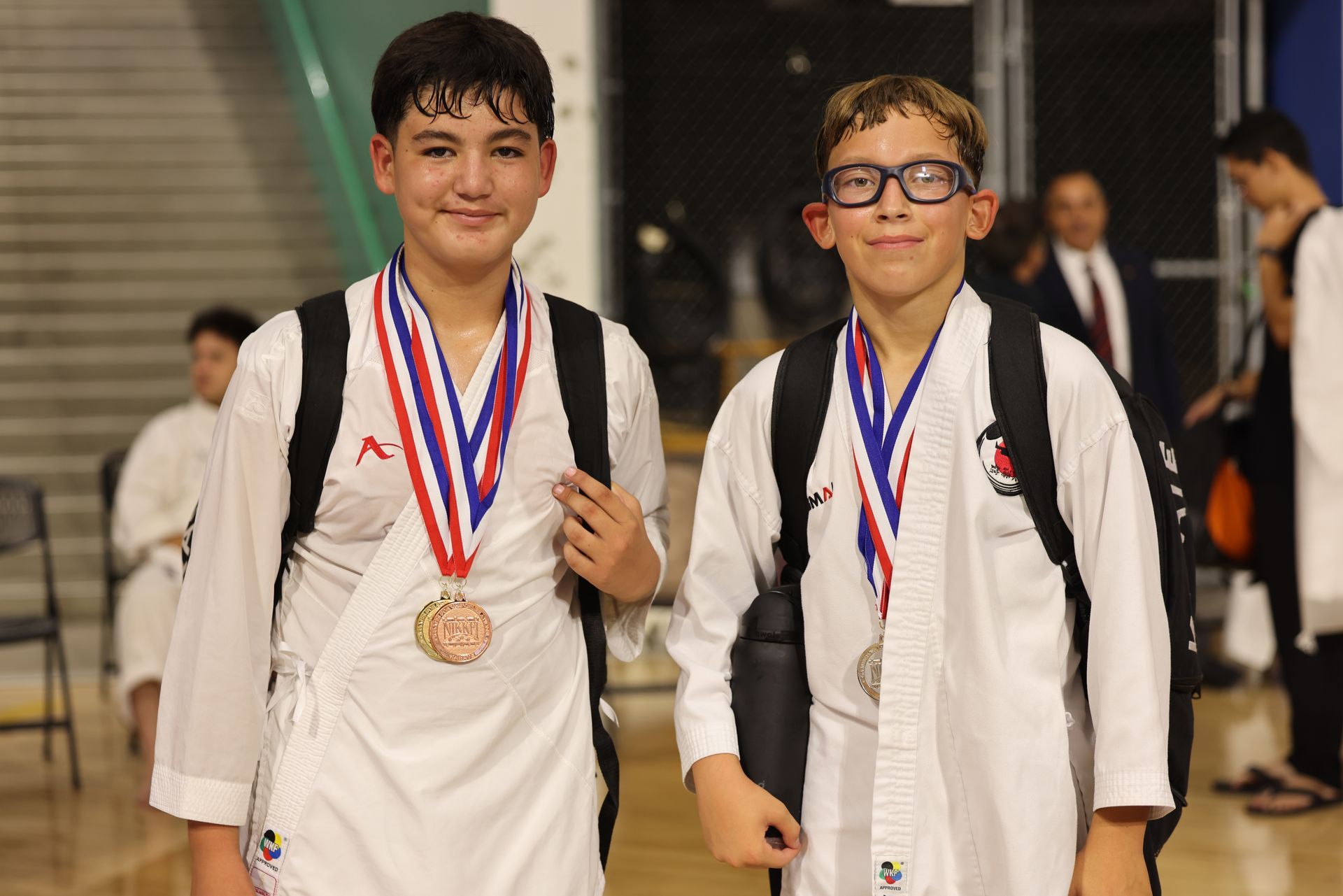 Two children in white martial arts uniforms with medals, standing indoors and smiling at the camera.