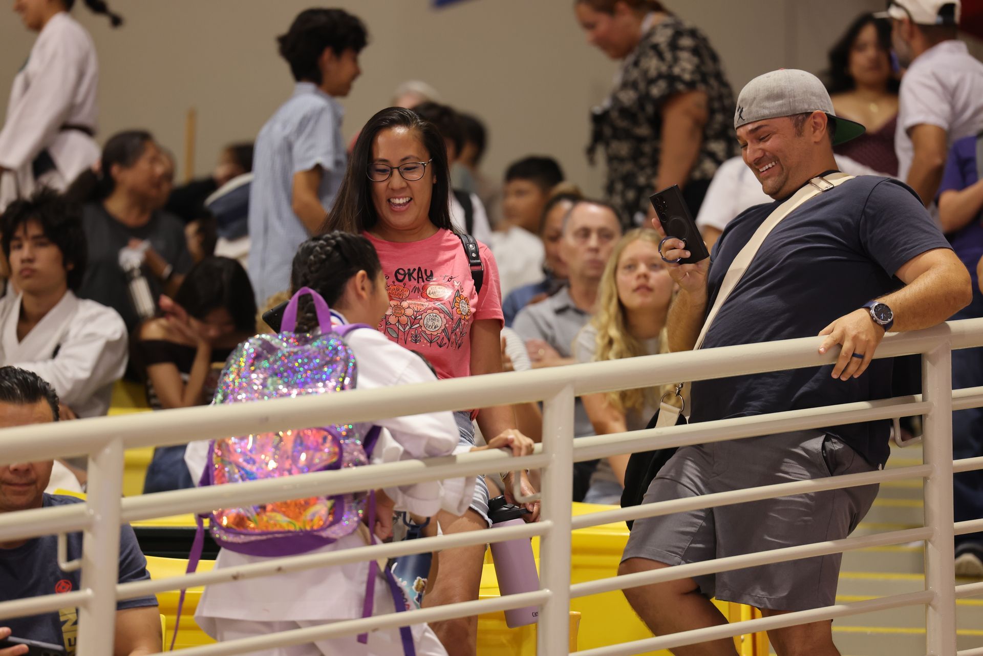 Crowd seated behind a railing, with a woman holding a shiny purple bag and a man leaning nearby.