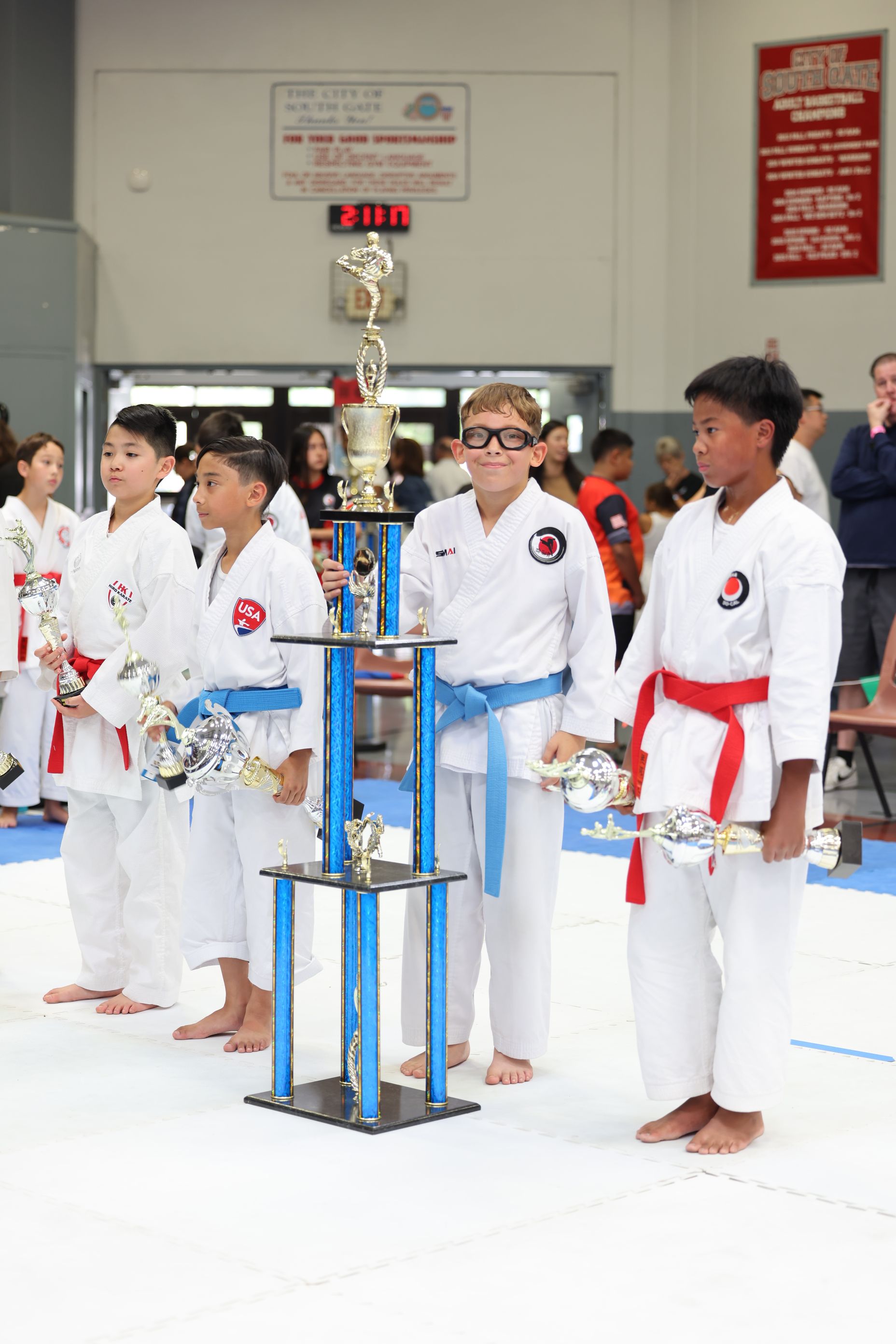 Karate tournament: Kids in white uniforms with trophies, a tall trophy stand, and spectators in a gymnasium.