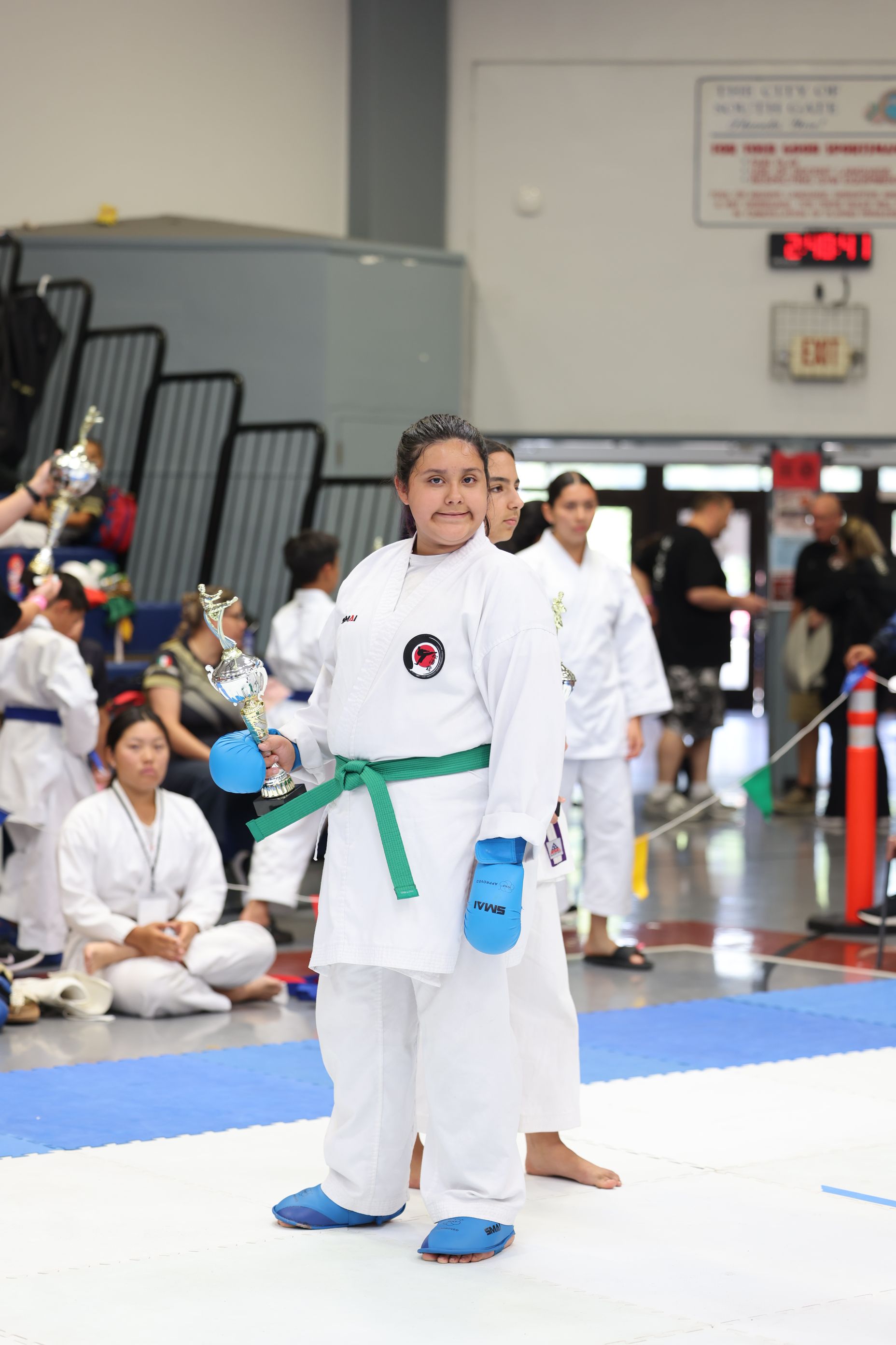 Young karateka in a white uniform with a green belt and blue gear, holding a trophy, smiles.