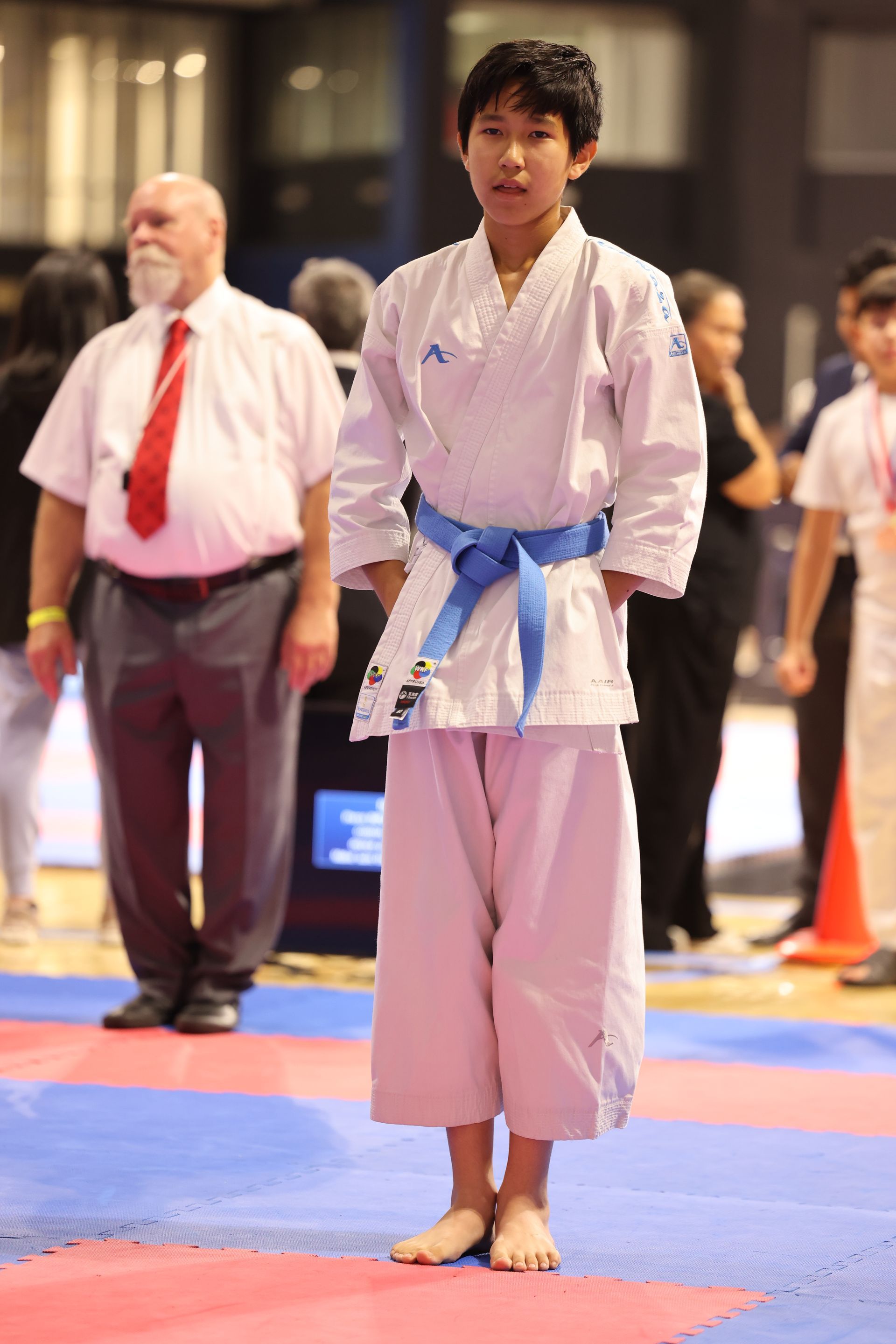 Karate competitor in a white gi with blue belt stands on a mat during a tournament.