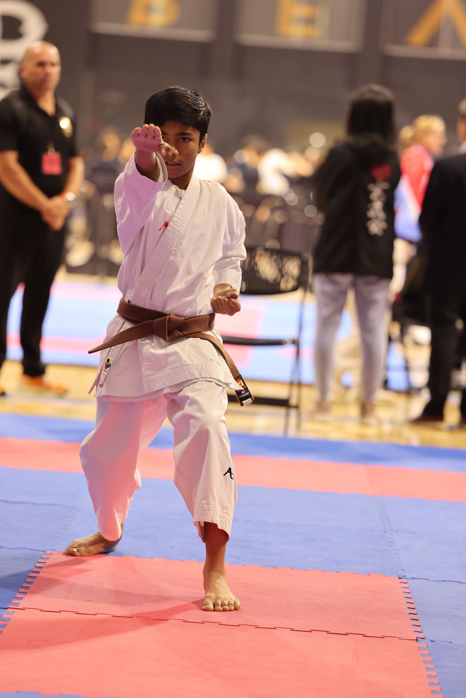 Karate competitor in a white gi and brown belt strikes a pose on a red-and-blue mat at an indoor tournament